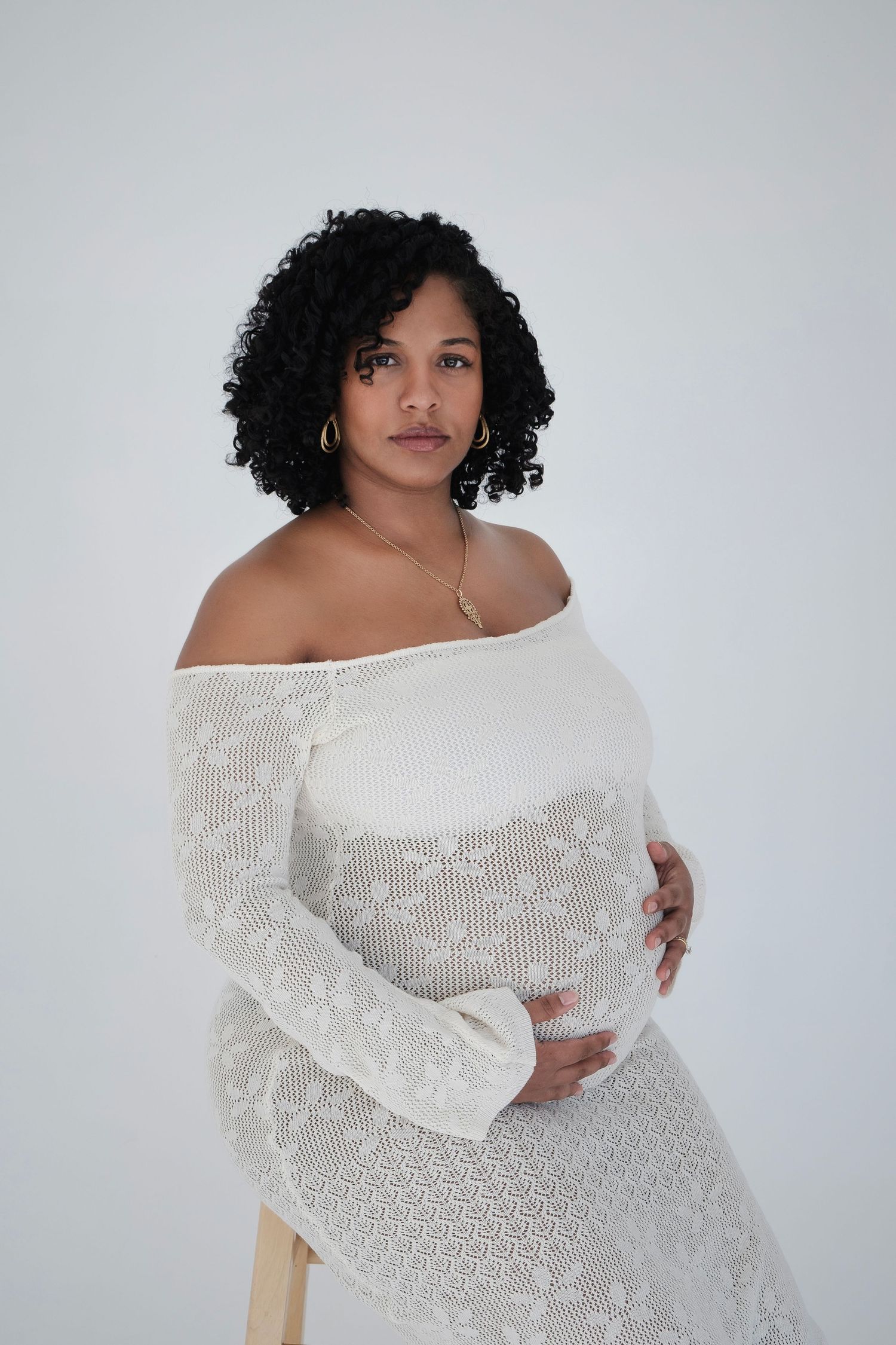 Expectant person in white lace off-shoulder dress poses against light background during maternity photoshoot.