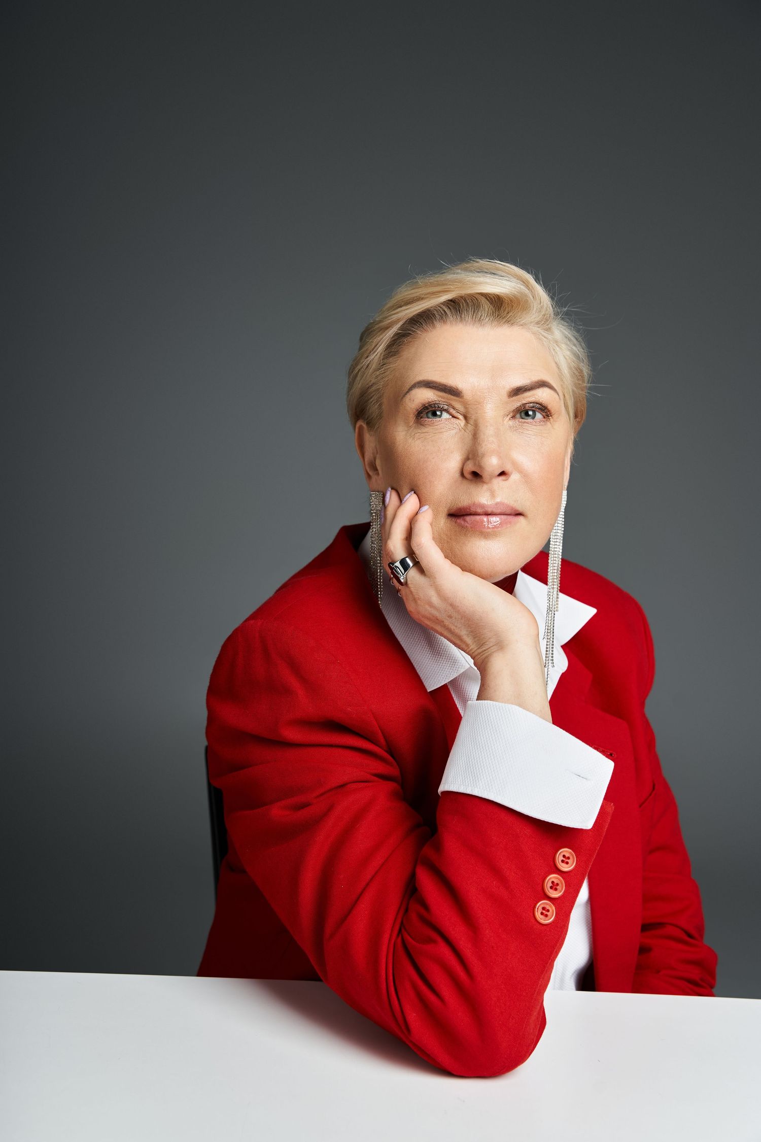 Person in bright red blazer resting chin on hand against grey background.