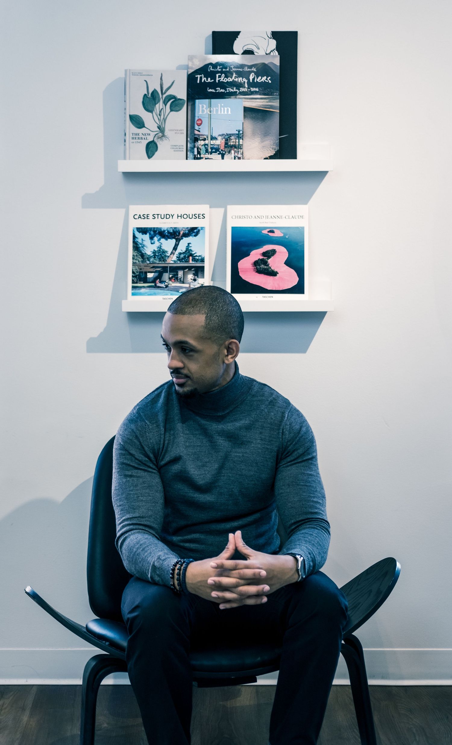 A person in a dark sweater sits thoughtfully on a chair against a white wall with floating shelves displaying artwork.