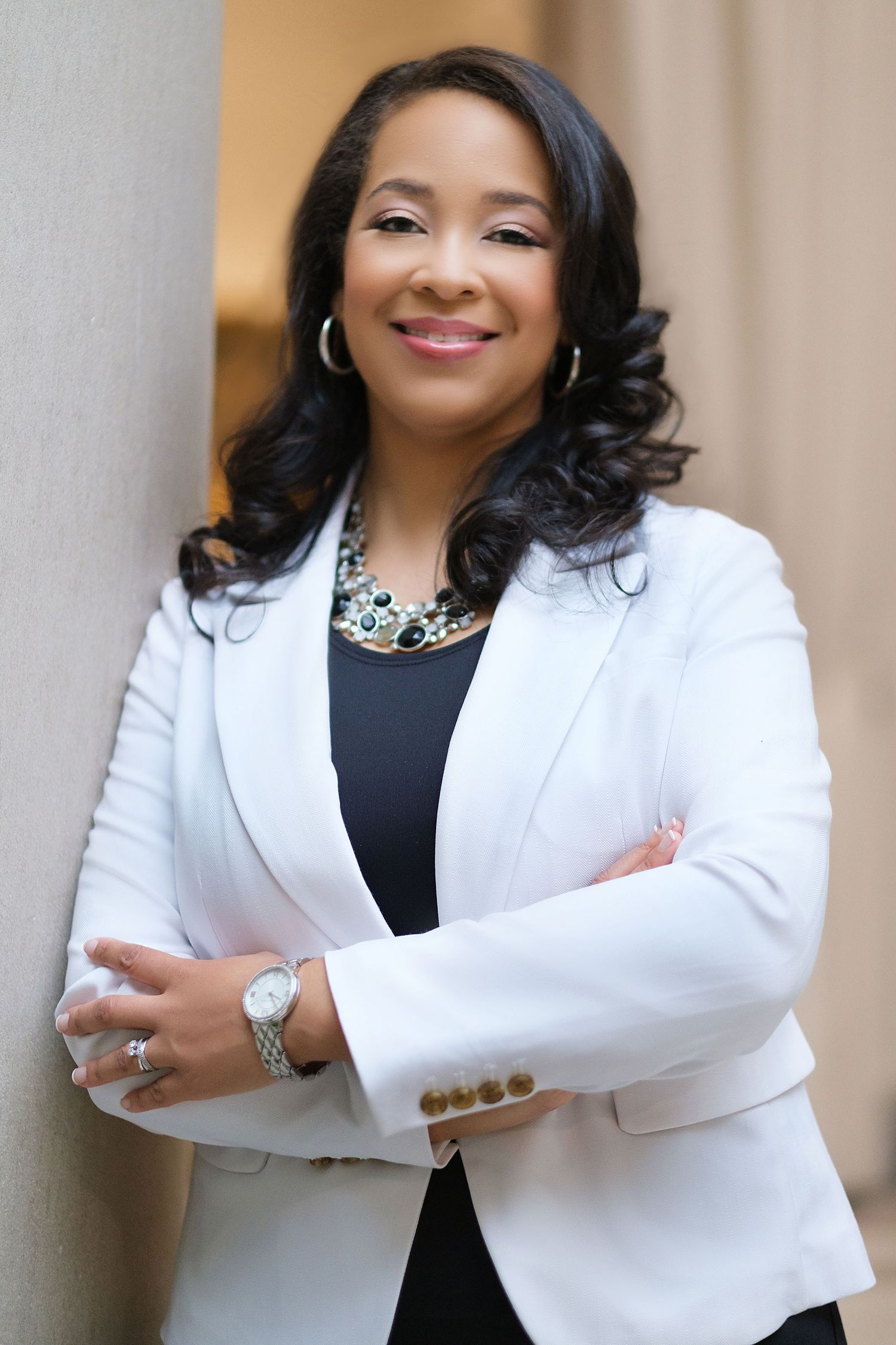 Professional portrait of a person in a white blazer and statement necklace standing with arms crossed against neutral background.