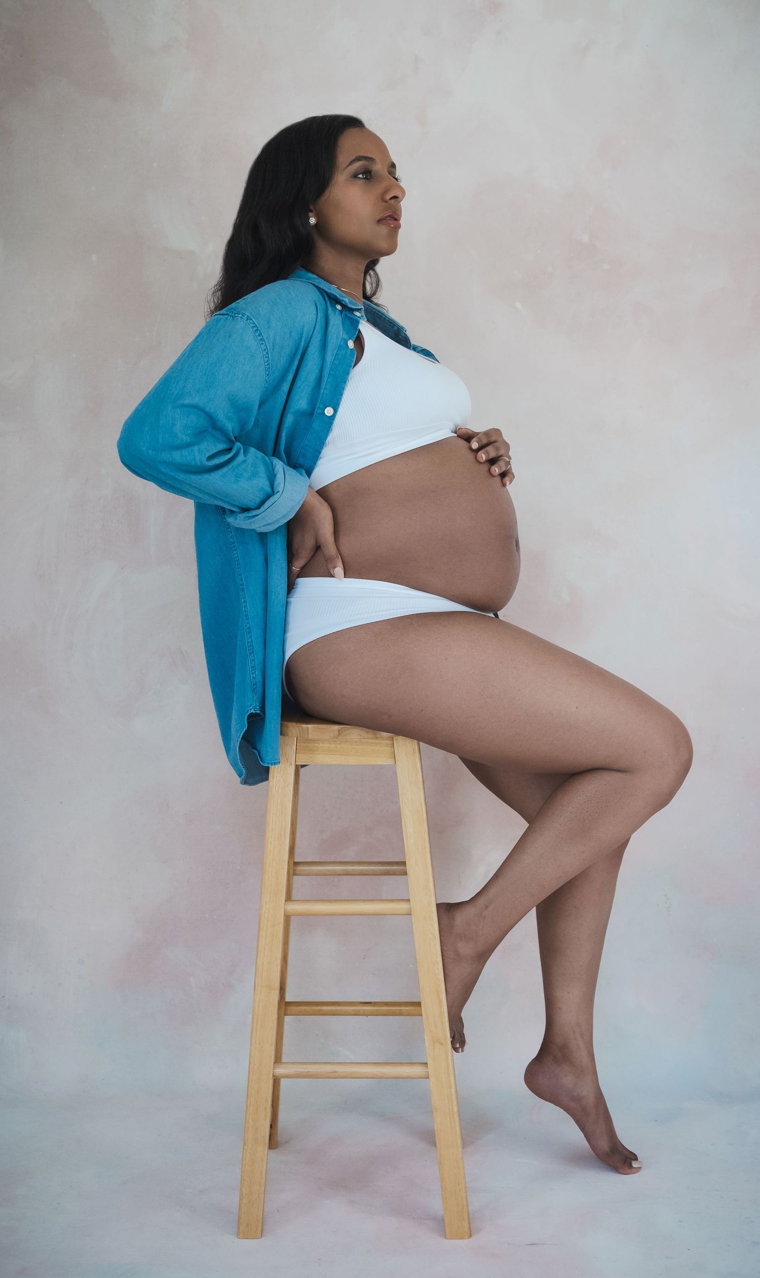 A pregnant figure in white undergarments and turquoise cardigan poses elegantly on a wooden stool against a neutral backdrop.