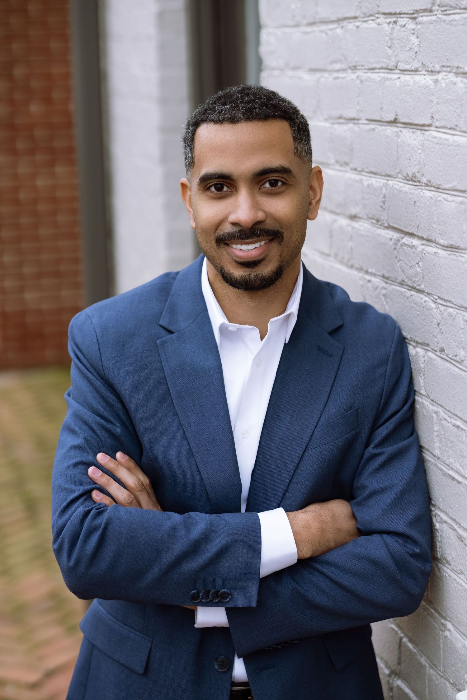 Professional headshot of someone in a navy blazer and white shirt smiling while standing against a white brick wall.