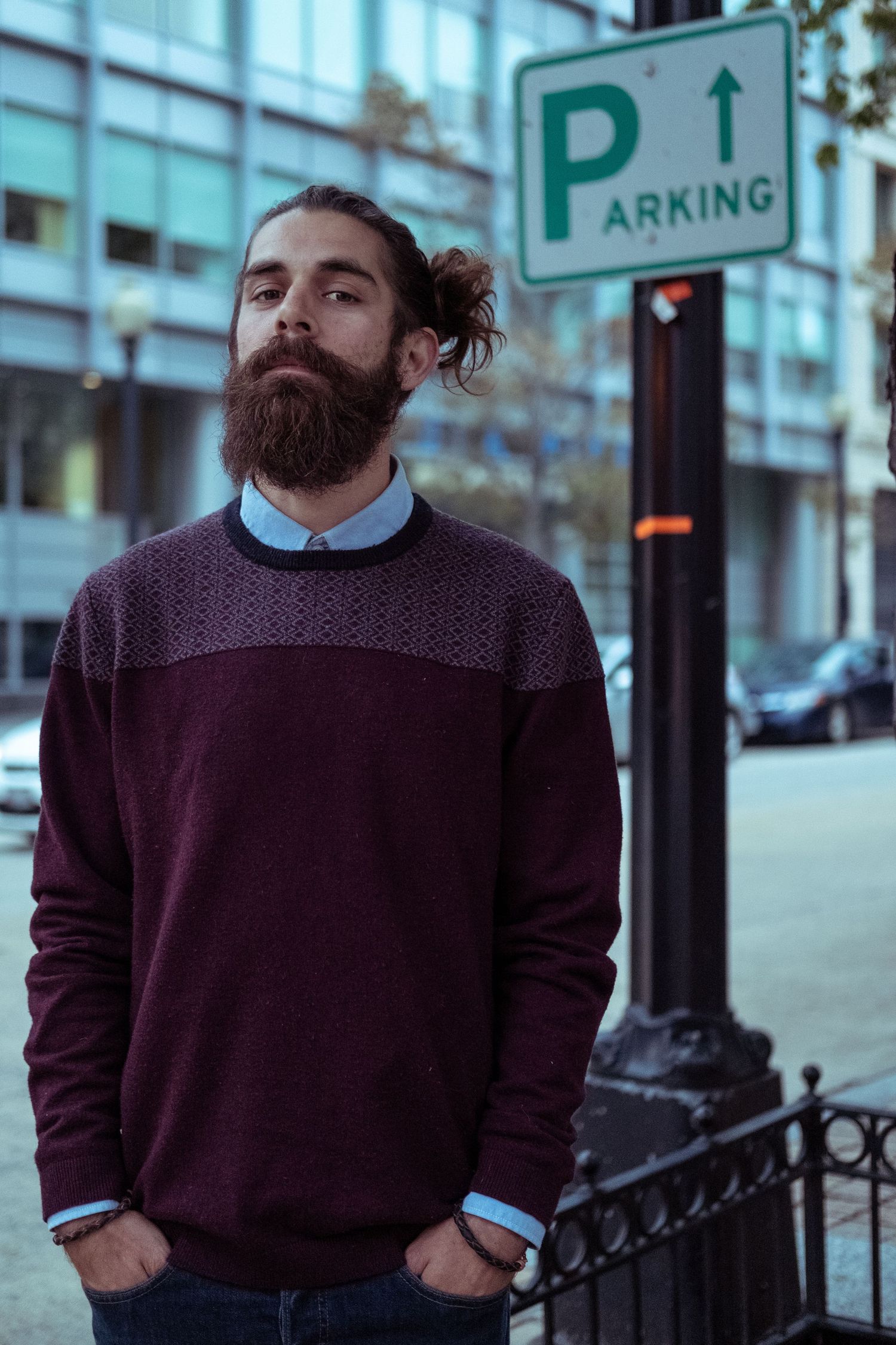 Person in burgundy sweater and light blue collared shirt stands on urban street near parking sign.