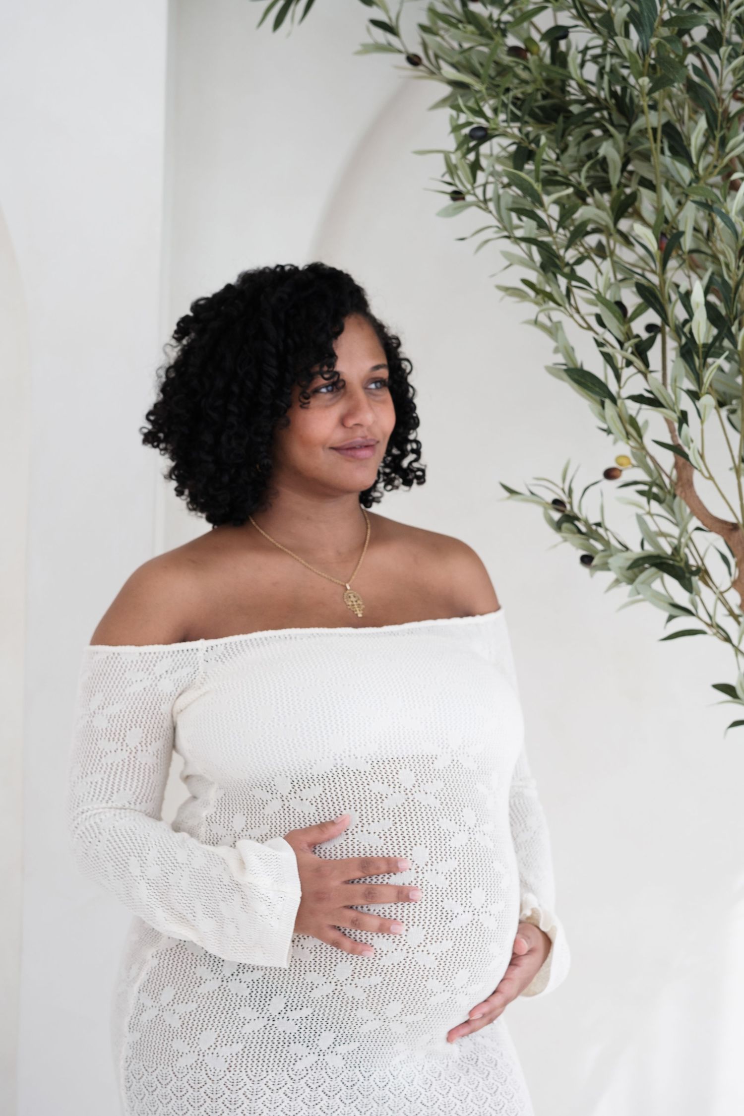 An expectant mother in a white off-shoulder dress poses against a white wall with olive branches.