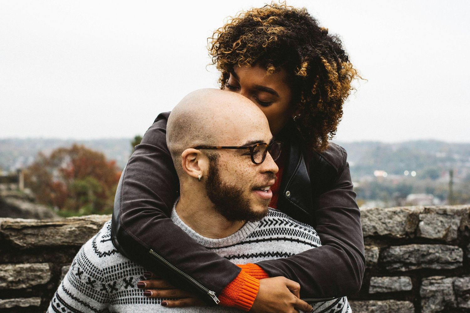 A couple shares a tender embrace while sitting on a stone wall with a scenic backdrop.