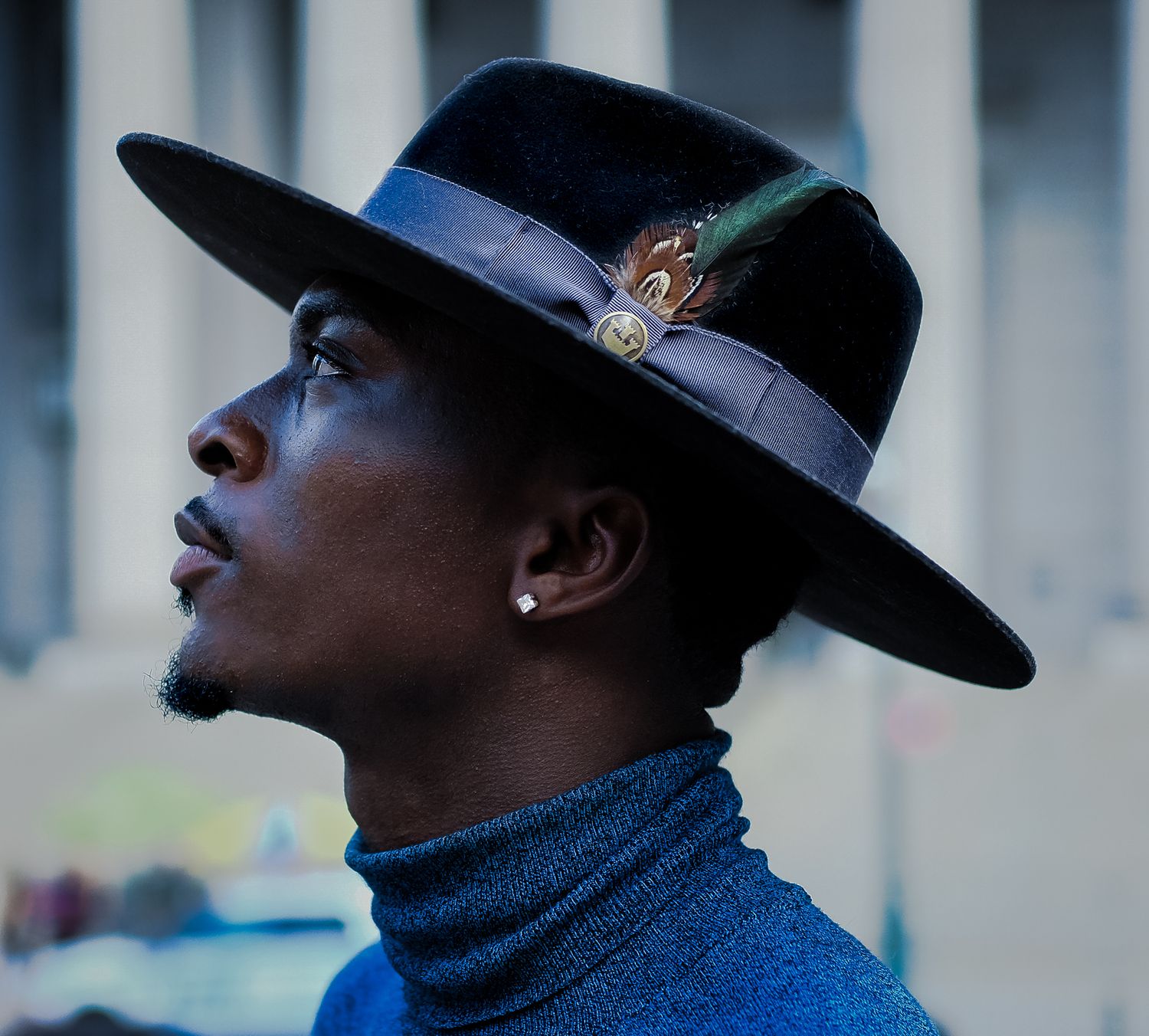 A person in a blue turtleneck and black hat gazes upward against a blurred urban background.