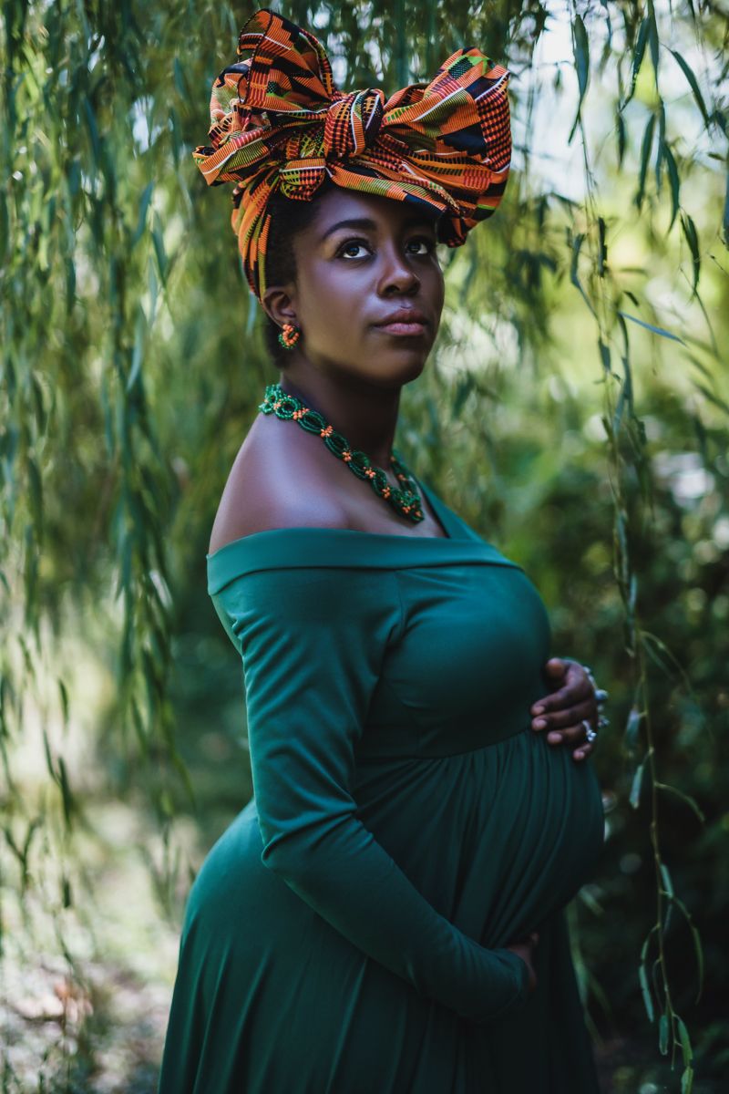 A pregnant woman in a green dress and orange headwrap poses gracefully under hanging willow tree branches.