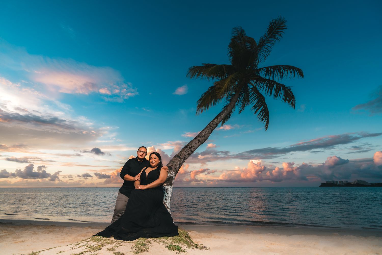 Couple in black outfits poses under a leaning palm tree on a tropical beach at sunset.