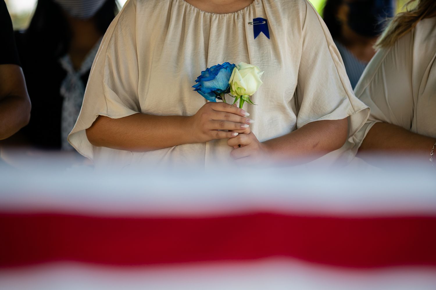 Guam funeral photographer capturing a woman in beige dress holding blue and white flowers stands before an American flag-draped casket.
