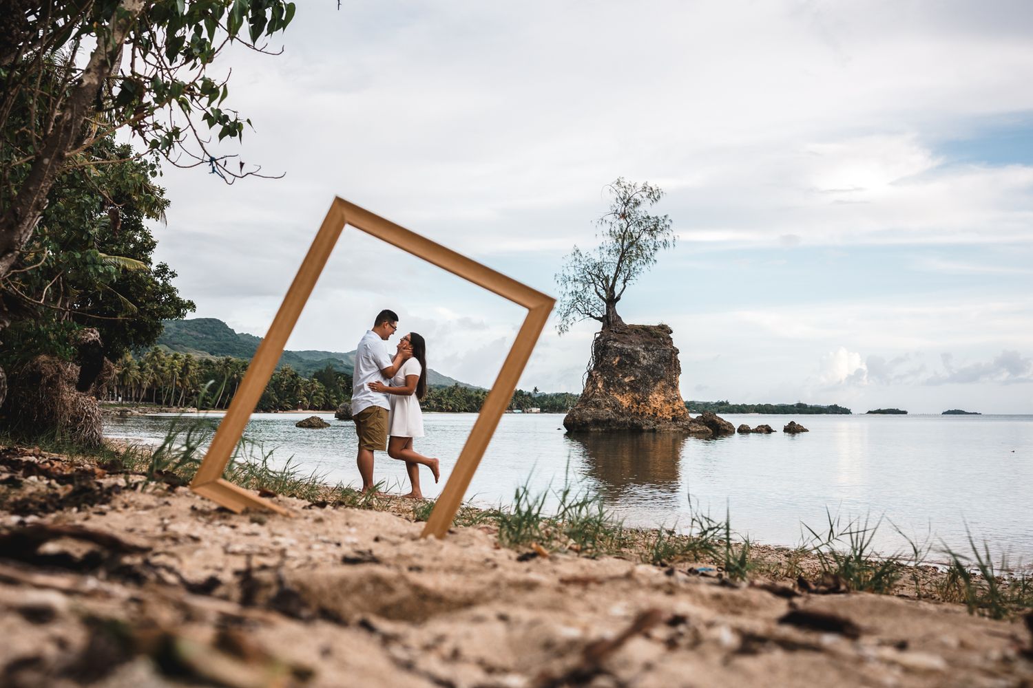 Couple embracing inside a wooden frame on a tropical beach shore with rocky outcrops and calm ocean waters.
