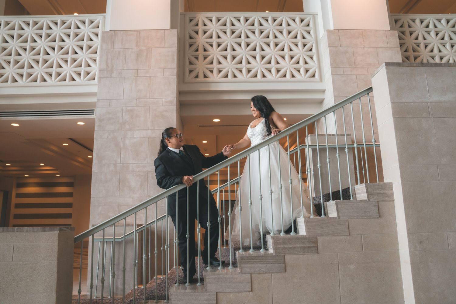 Bride and groom walking up elegant staircase in a grand venue, bride in white gown, groom in black suit.