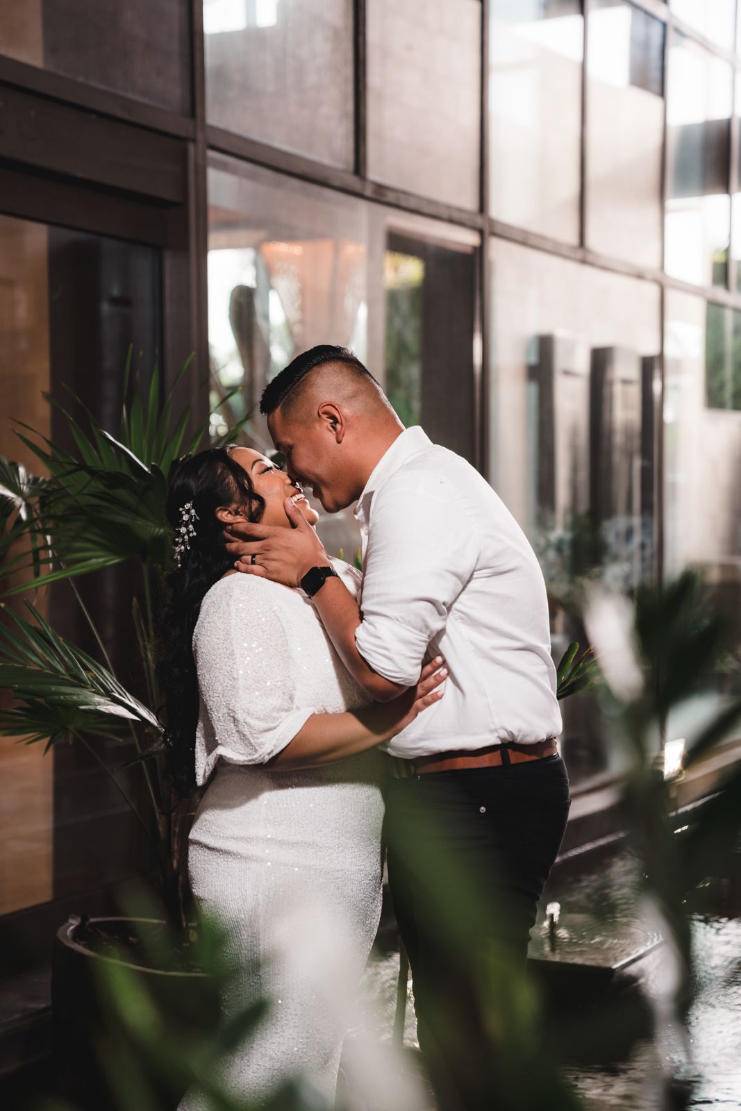 Couple sharing a romantic kiss surrounded by lush tropical plants near a modern glass building at night.