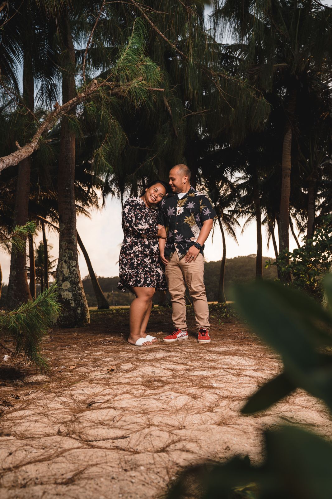 A couple laughs together among tropical palm trees on a sandy path, the woman in a floral dress and man in a Hawaiian shirt.