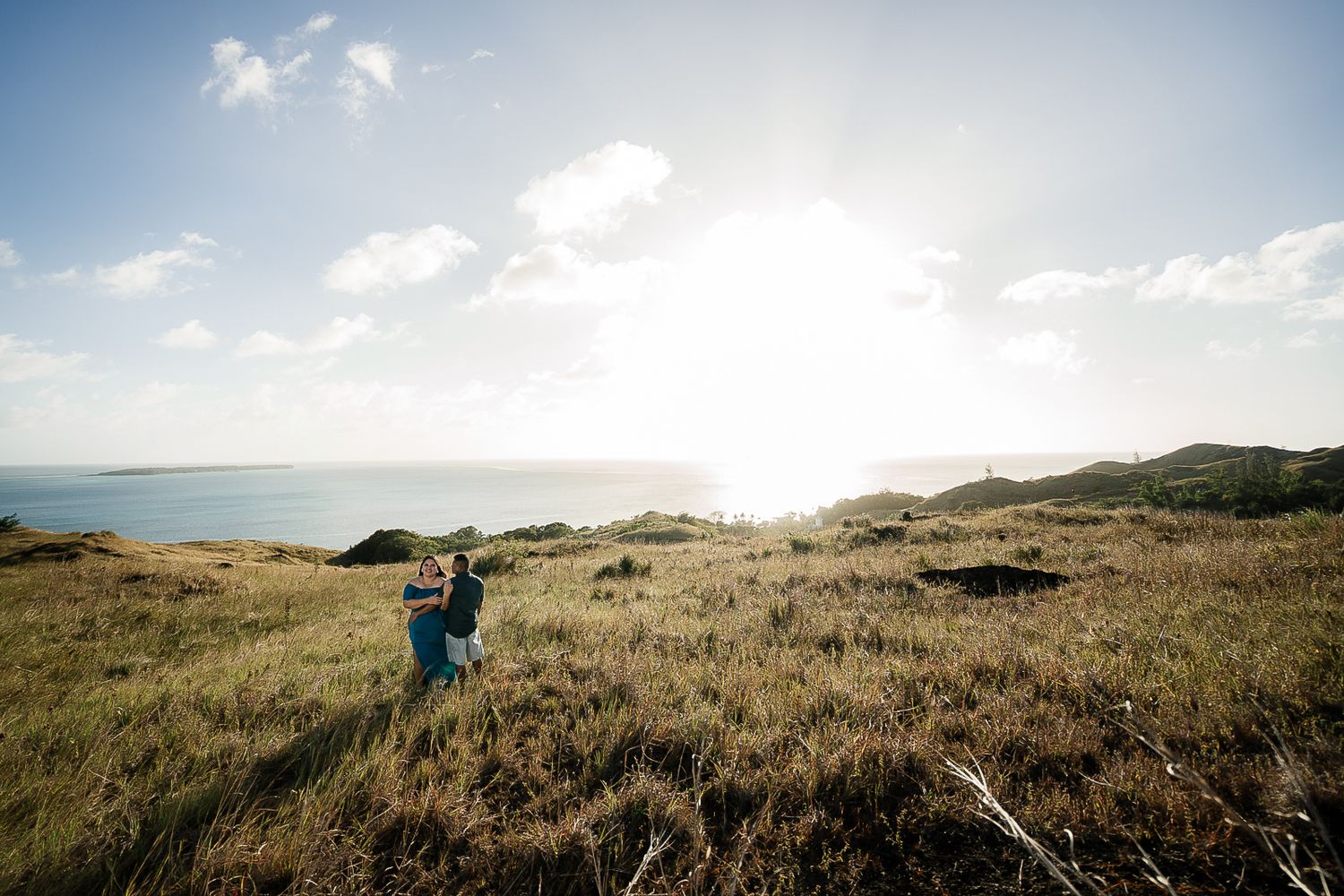 Couple embracing in golden coastal grasslands with ocean horizon and bright sun in background.