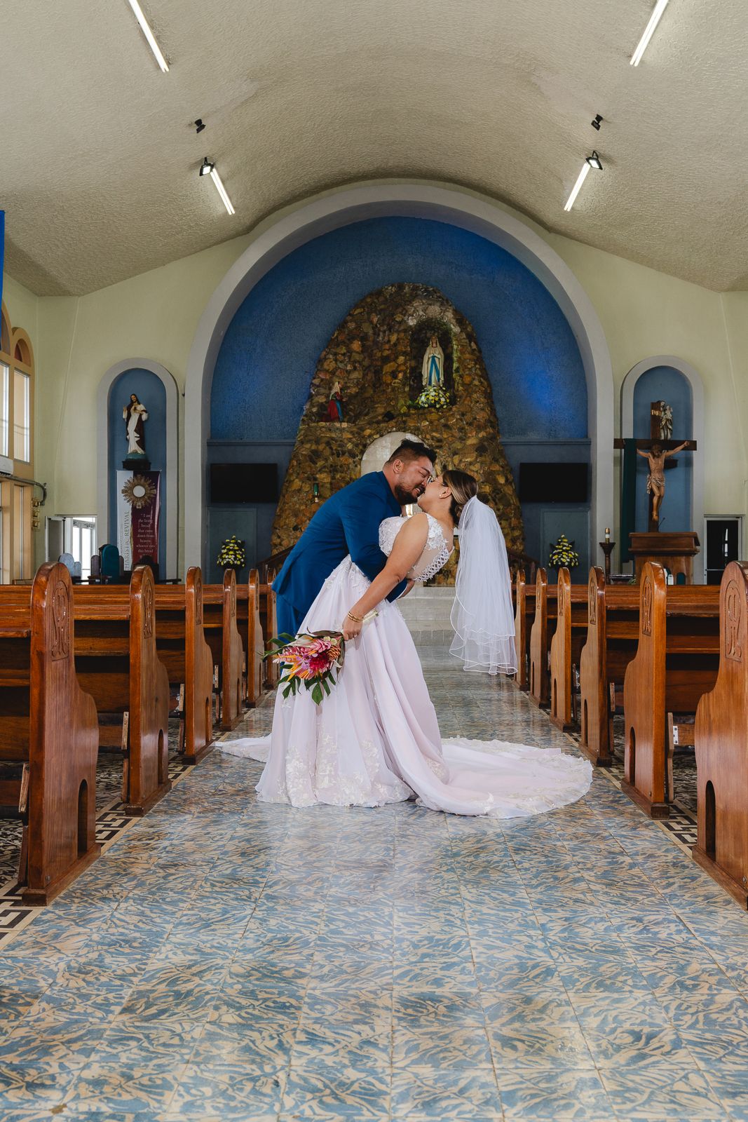 Guam wedding photographer capturing the bride and groom sharing a dip kiss in church aisle with blue arched altar backdrop.