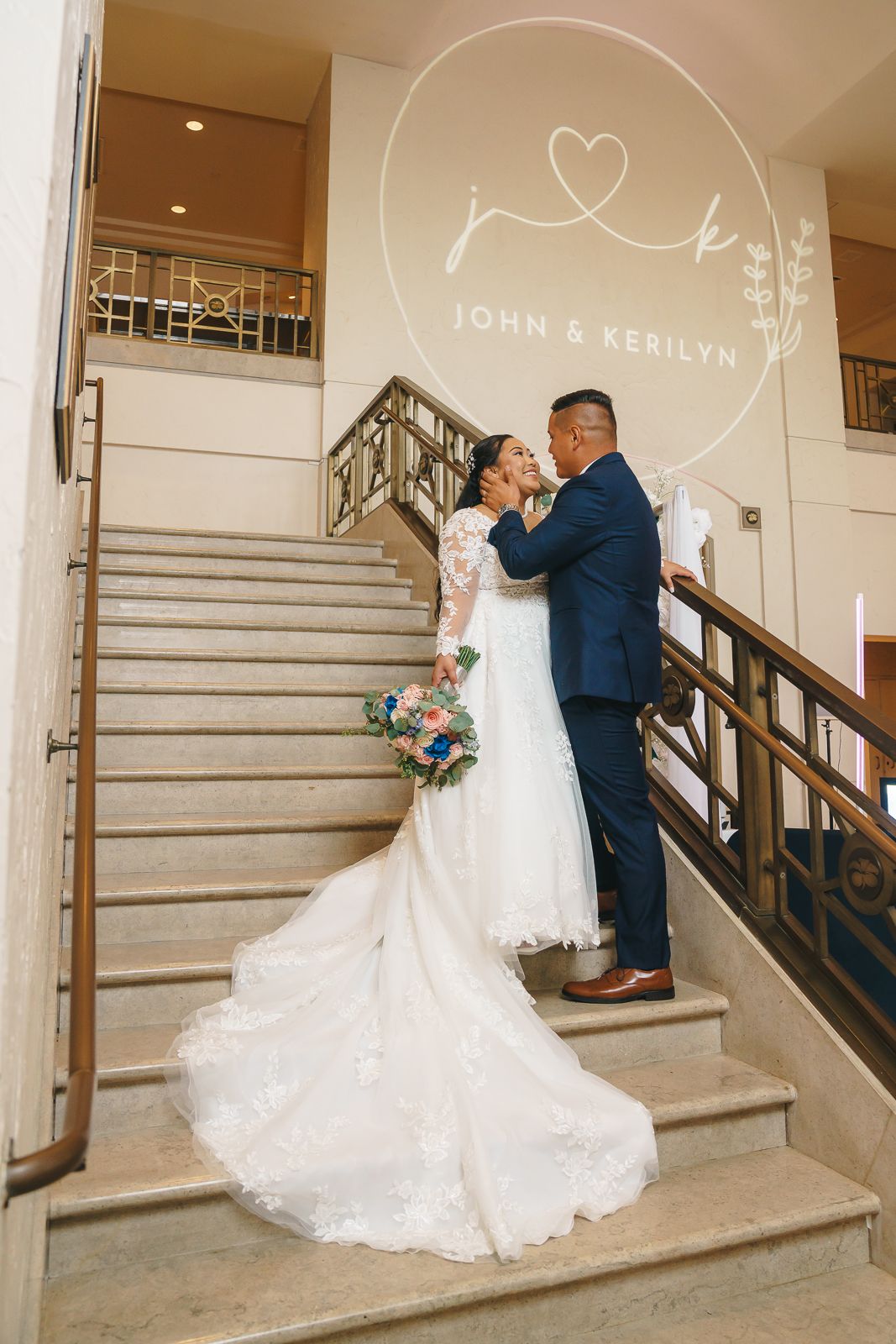 Guam wedding photographer capturing the bride and groom's embrace on grand staircase beneath John and Kerilyn wedding monogram sign.