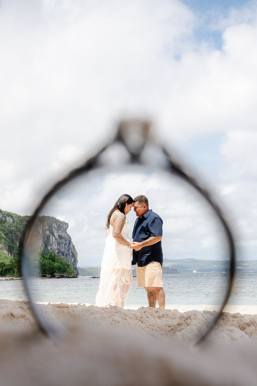 Couple embracing on a tropical beach, framed by a wedding ring in the foreground.