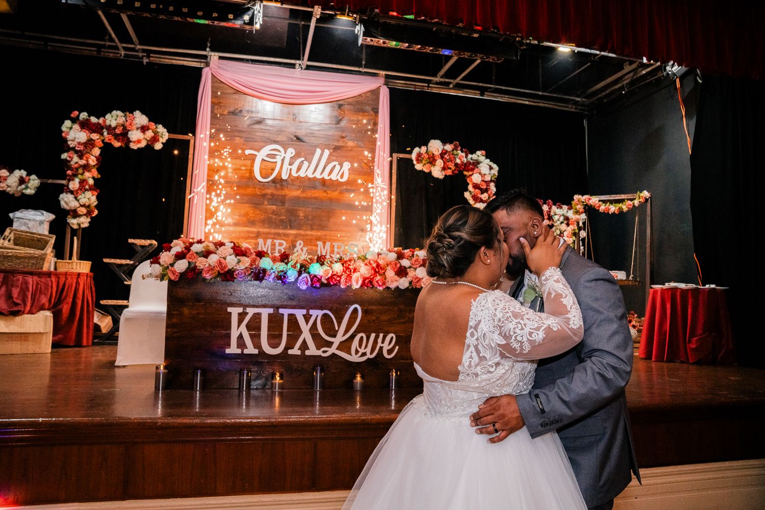 Guam wedding photographer capturing the bride and groom sharing a kiss at wedding reception with floral backdrop and KUXLove sign.