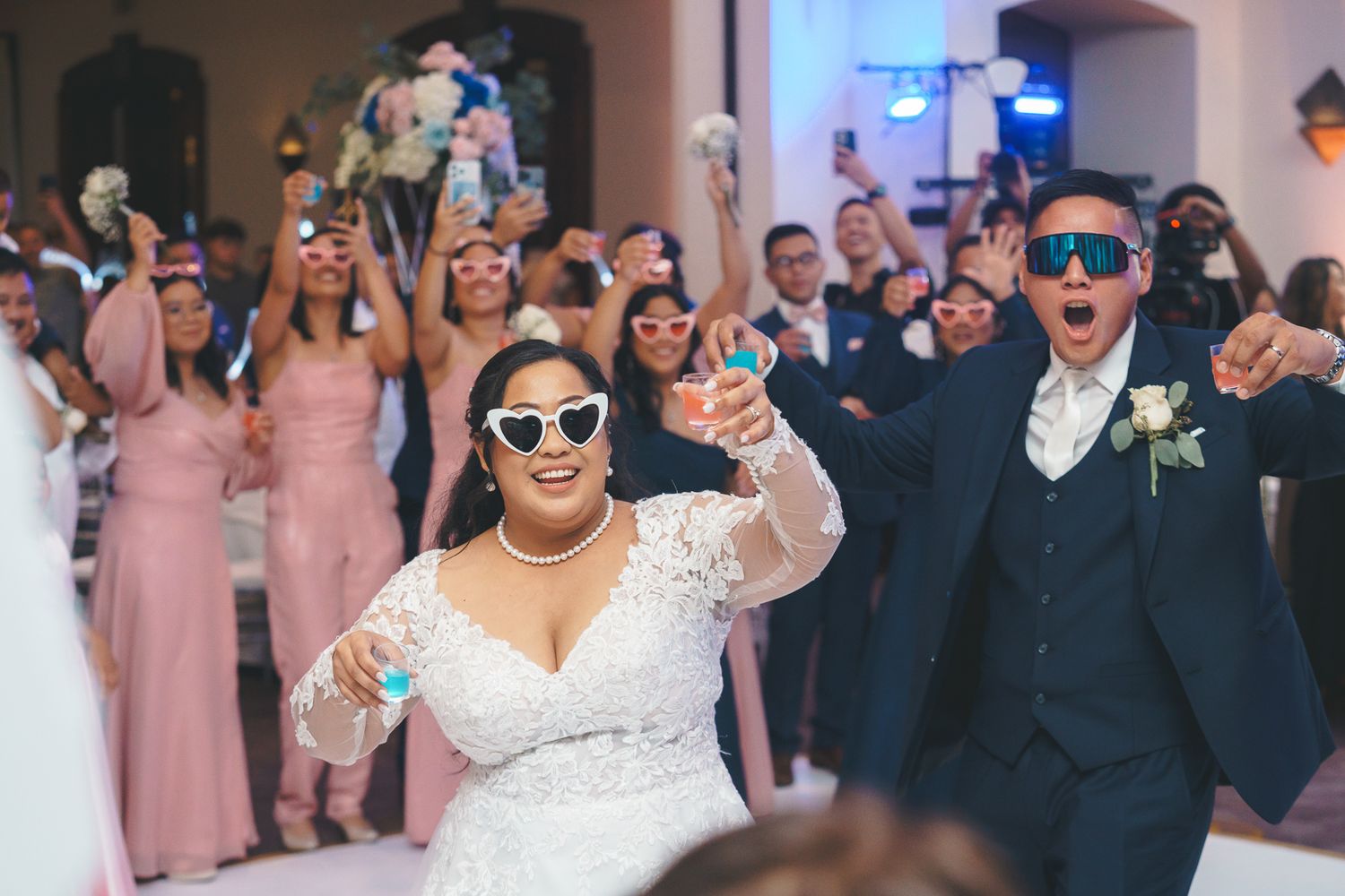 Bride and groom wearing sunglasses celebrate joyfully at wedding reception with bridesmaids in pink dresses behind them.