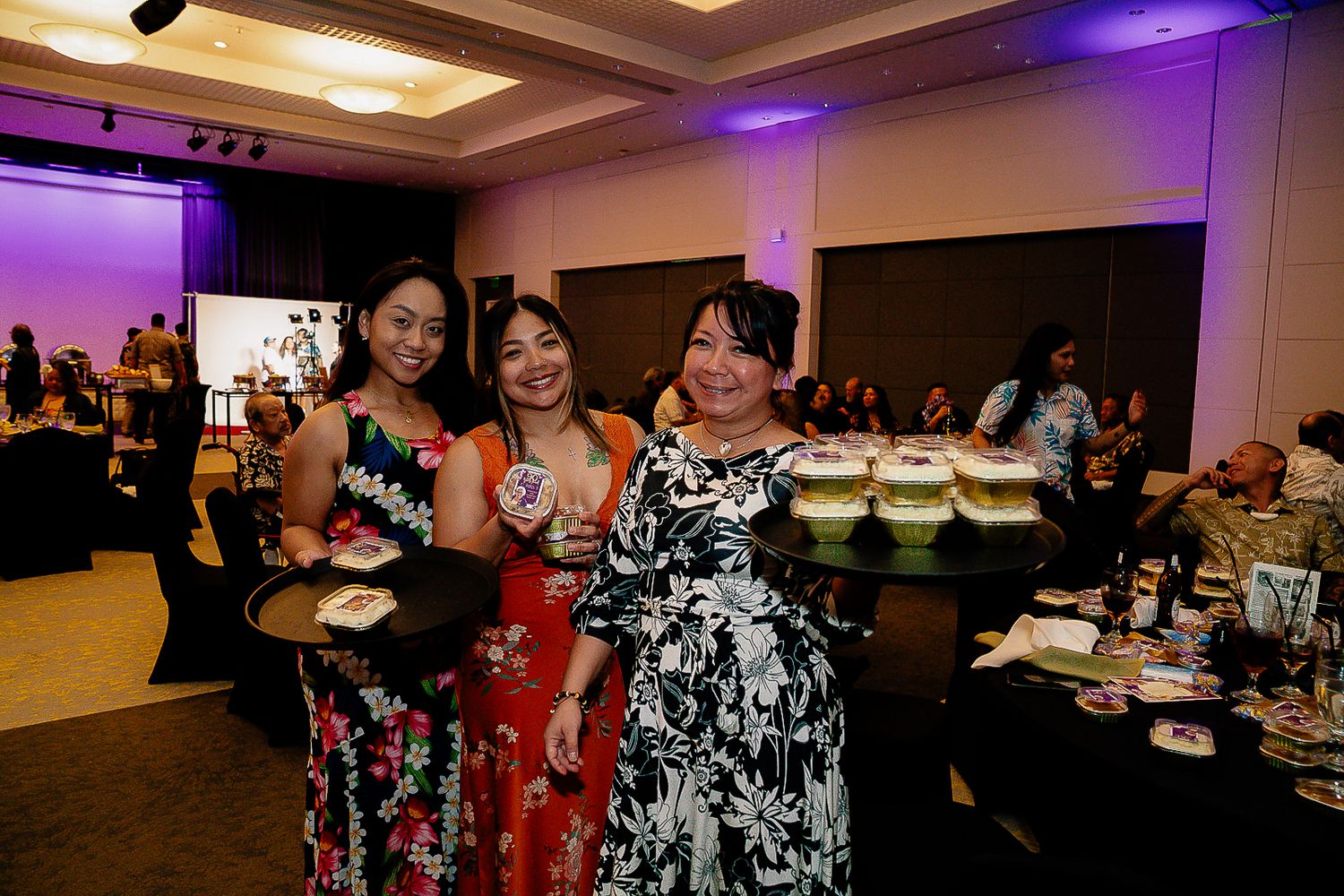 Three women in floral dresses holding food trays and smiling at a formal banquet event.