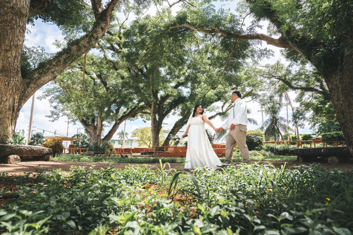 Guam wedding photographer capturing the bride and groom walk hand in hand through lush garden under arching trees.