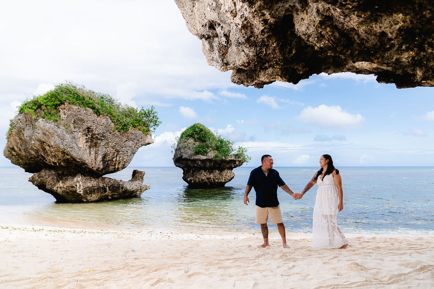 Couple holding hands on a tropical beach with dramatic rock formations and turquoise ocean in background.