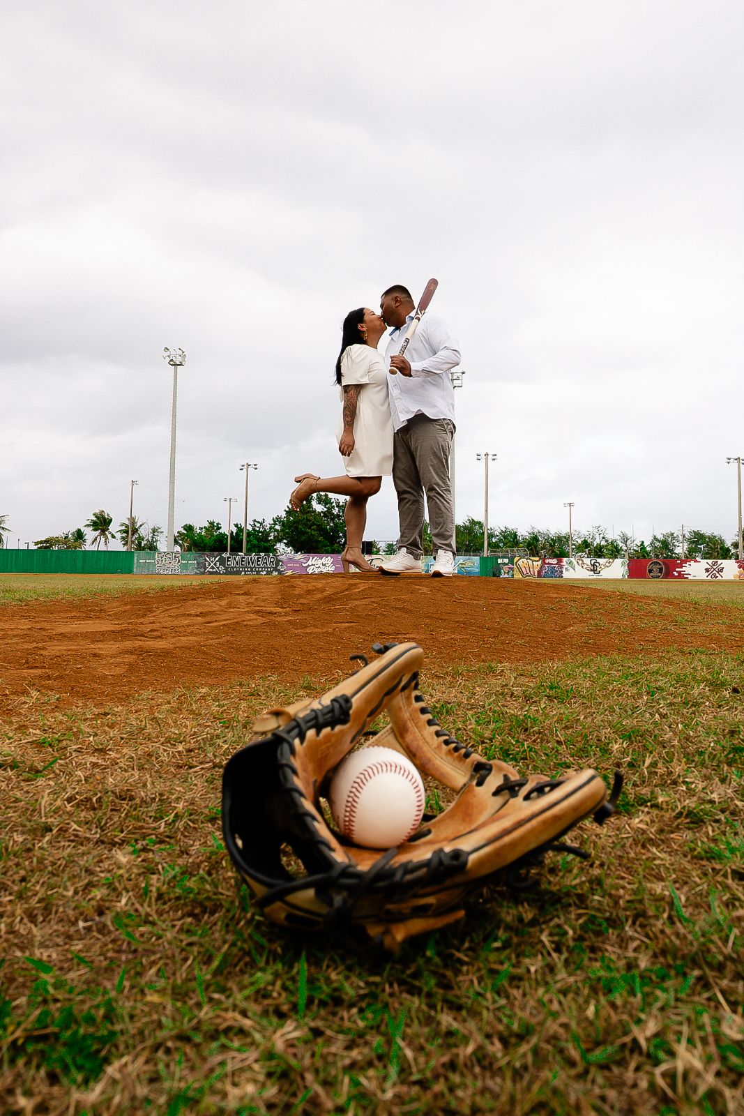 Couple sharing a romantic kiss on a baseball pitcher's mound with a glove and ball in the foreground.