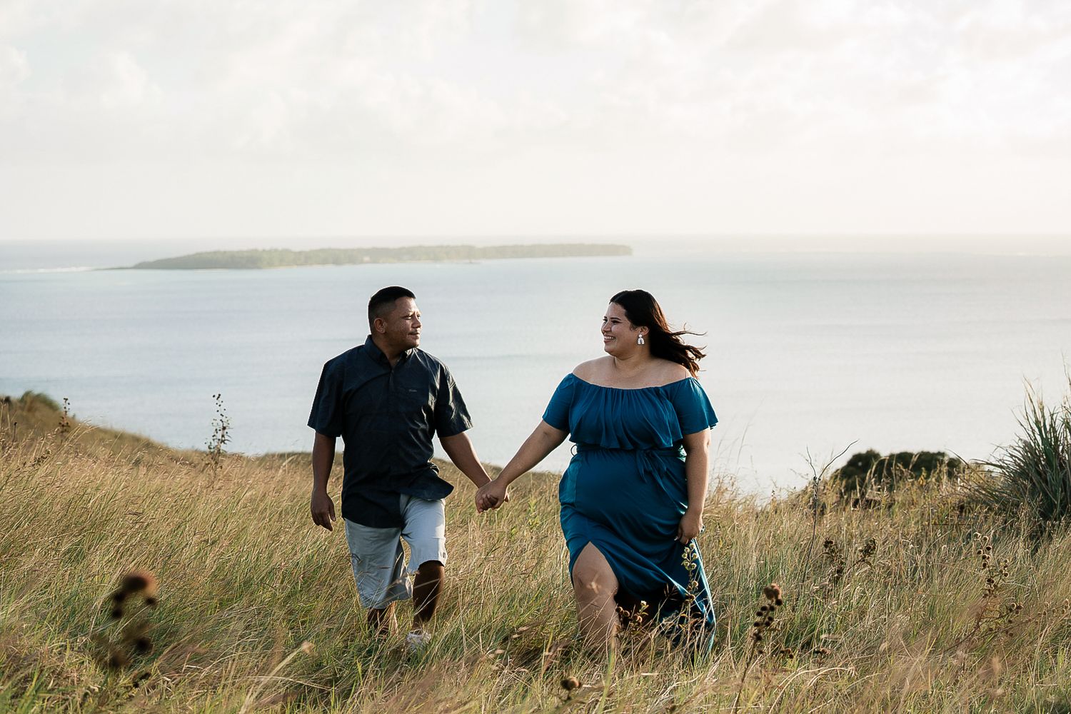 Couple holding hands walking through tall grass with ocean and island views in the background.