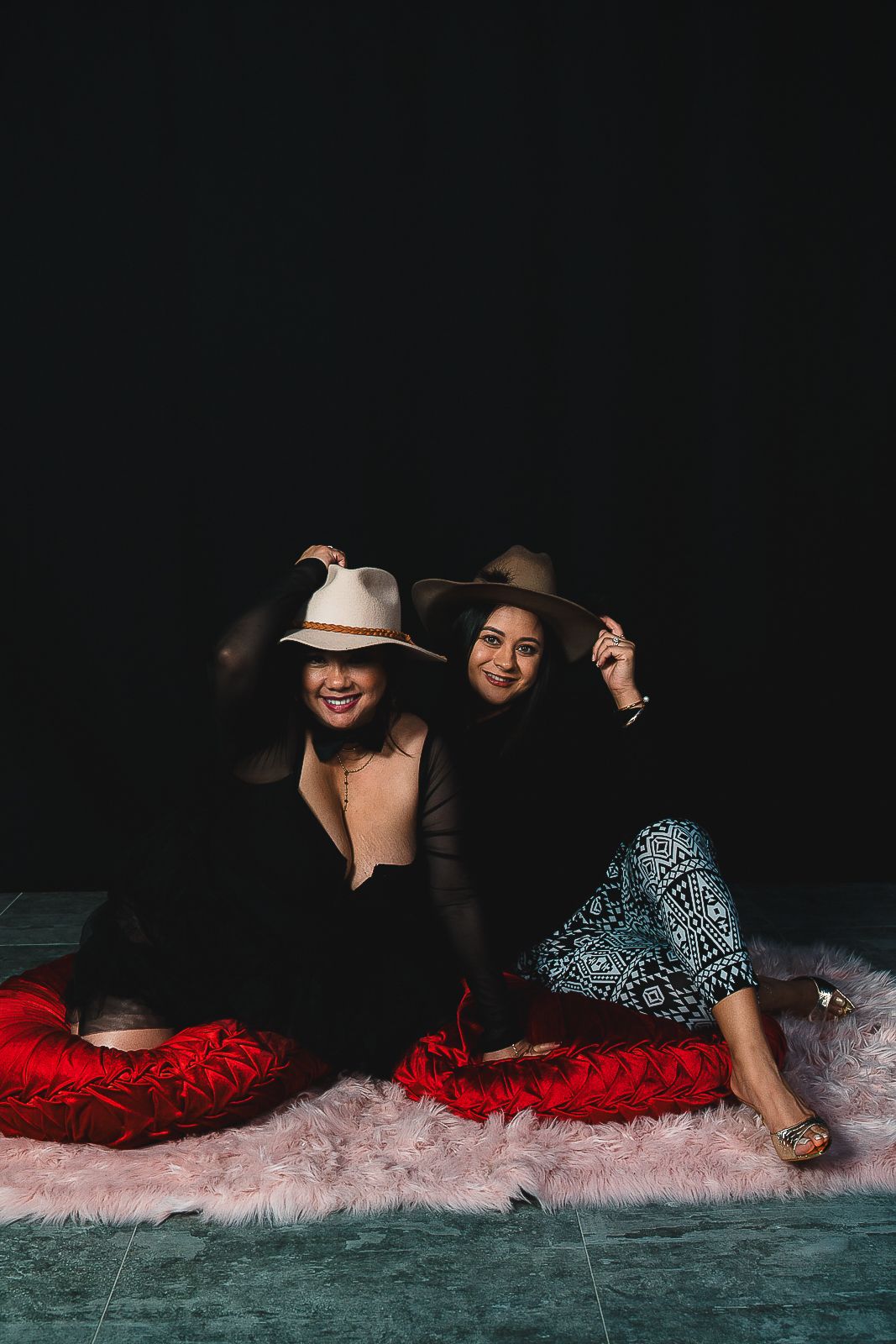Guam event photographer capturing two women sitting on colorful cushions wearing wide-brim hats against a dark background.