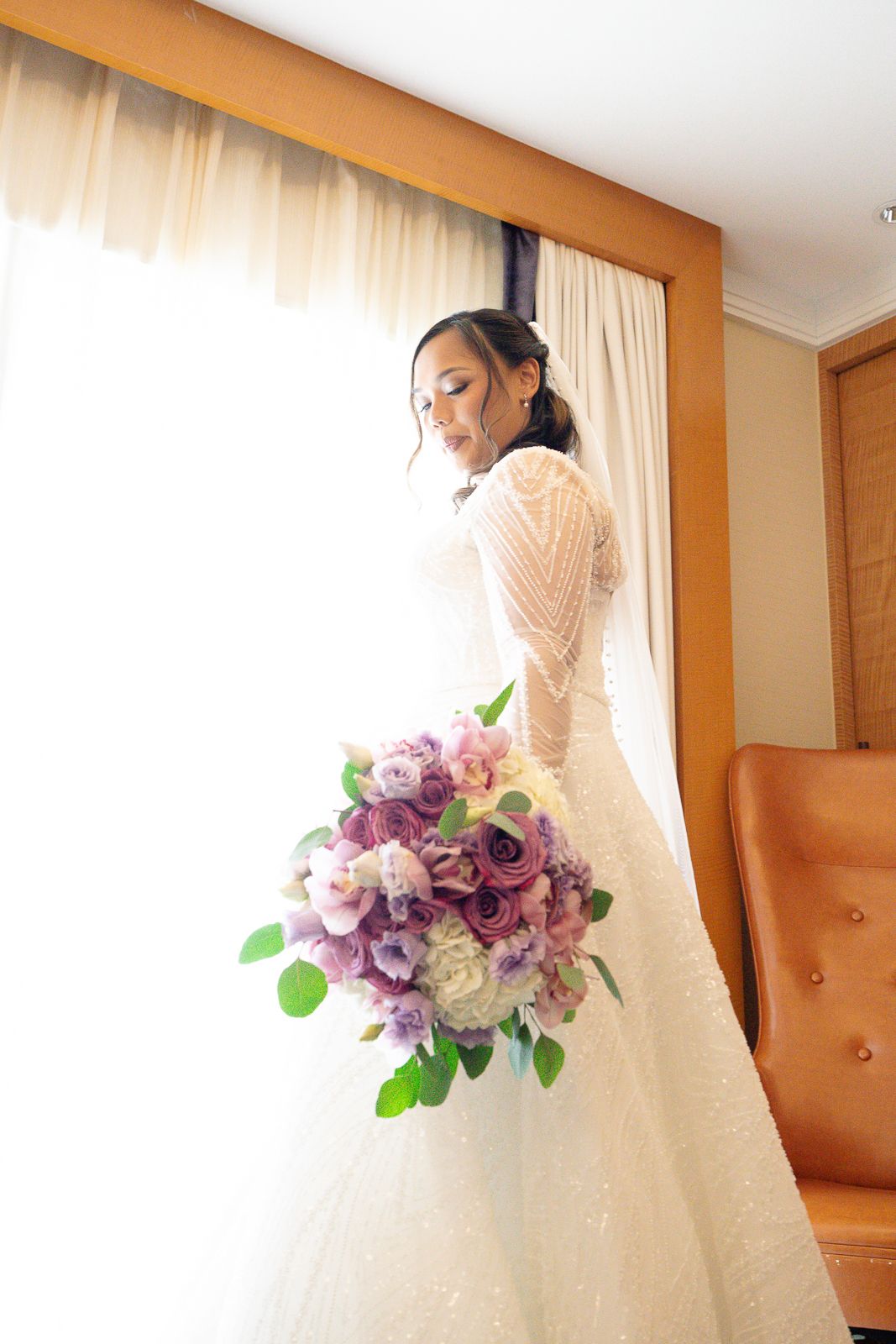 Guam wedding photographer capturing an elegant bride in lace gown holding purple and white floral bouquet by bright window.