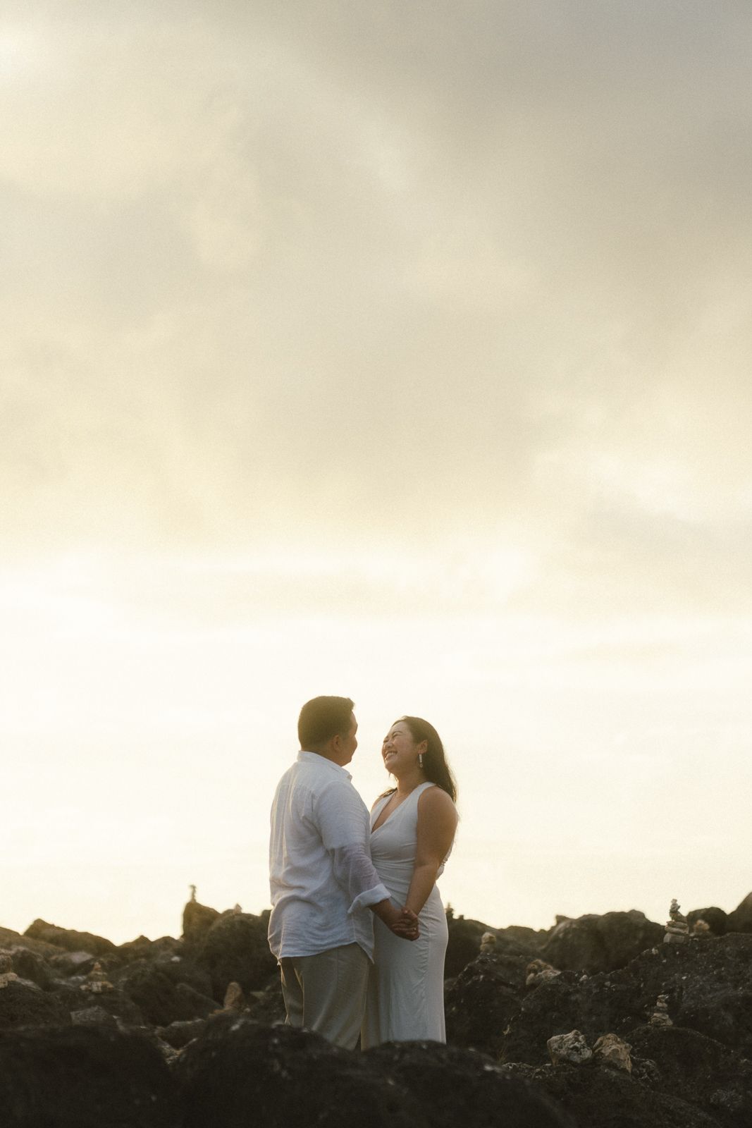 Couple holding hands on rocky shore at sunset, sharing a romantic moment against a dramatic cloudy sky.