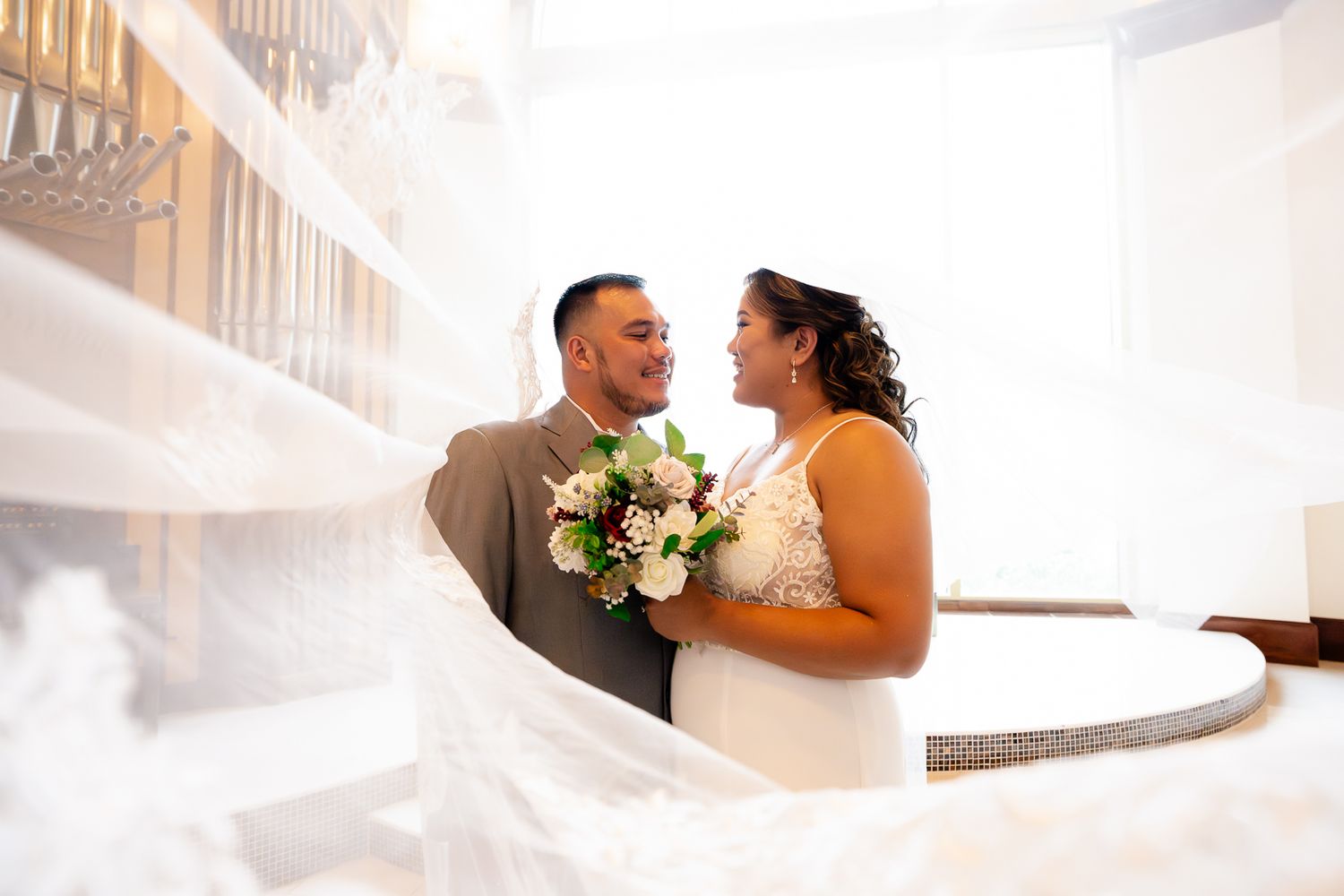 Guam wedding photographer capturing the bride and groom sharing a romantic moment indoors with bridal veil flowing in foreground.
