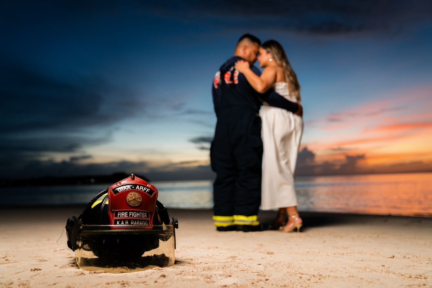 Couple embracing at sunset on beach with firefighter helmet in foreground during romantic engagement photo session.