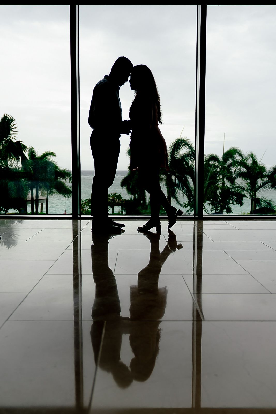 Romantic silhouette of couple nearly kissing reflected on glossy floor with palm trees and ocean view.