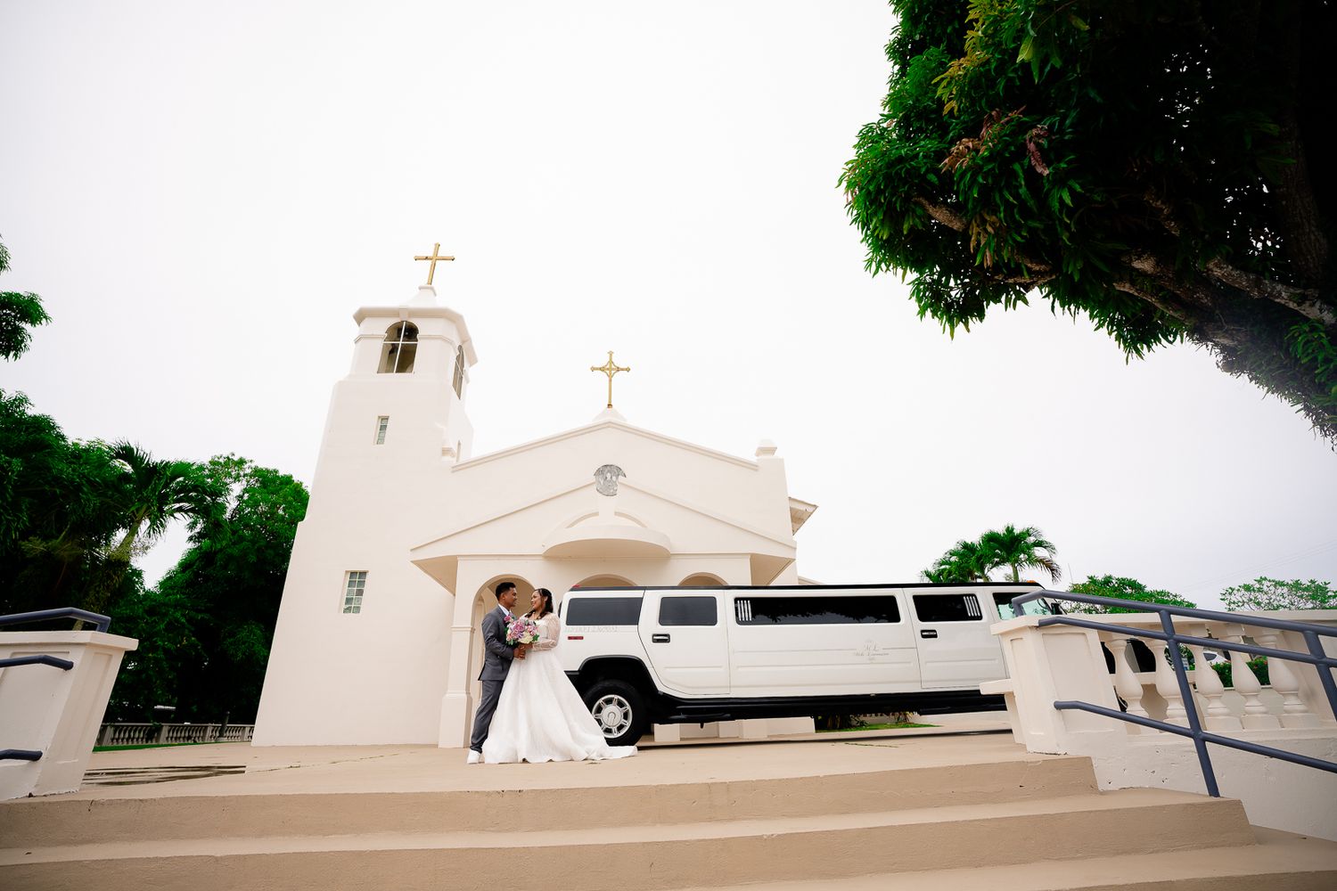 Guam wedding photographer capturing a wedding couple kissing in front of white church with stretch limo and tropical trees.