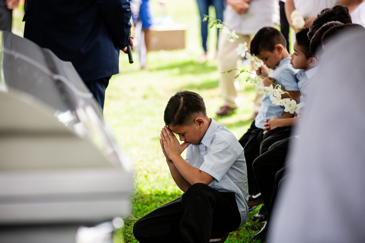 Guam funeral photographer capturing a young boy kneeling in prayer at a graveside funeral service beside a white casket outdoors.