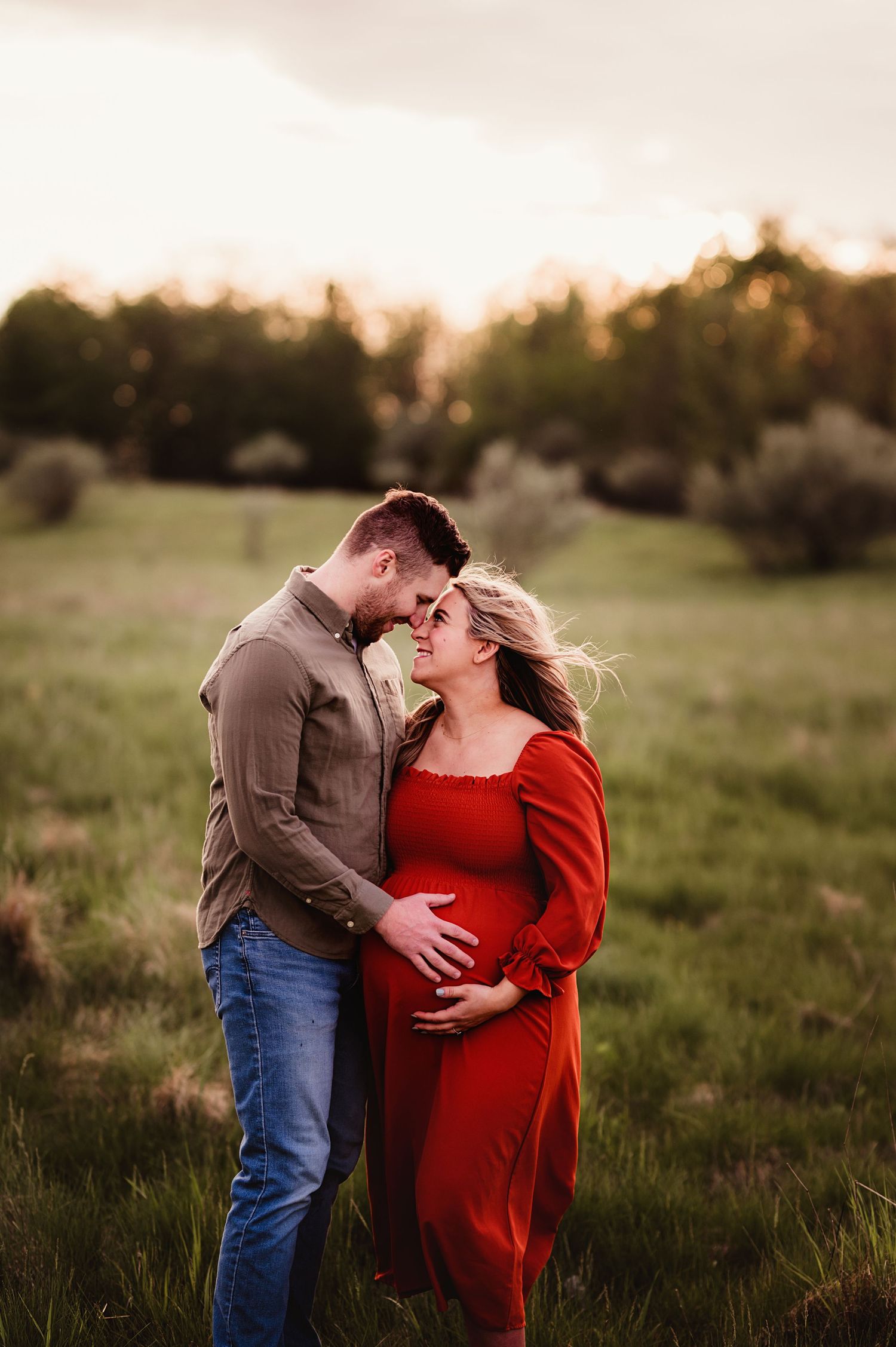 Romantic maternity photo of couple in field at sunset with mother wearing flowing red dress.