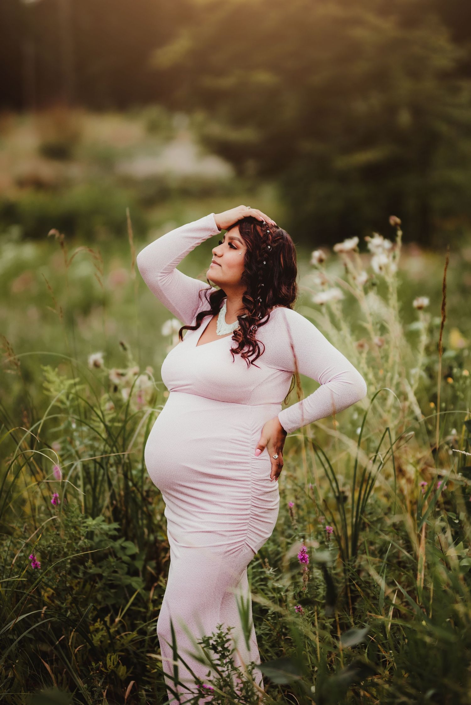 Maternity photo of woman in pink dress standing in meadow during golden hour.