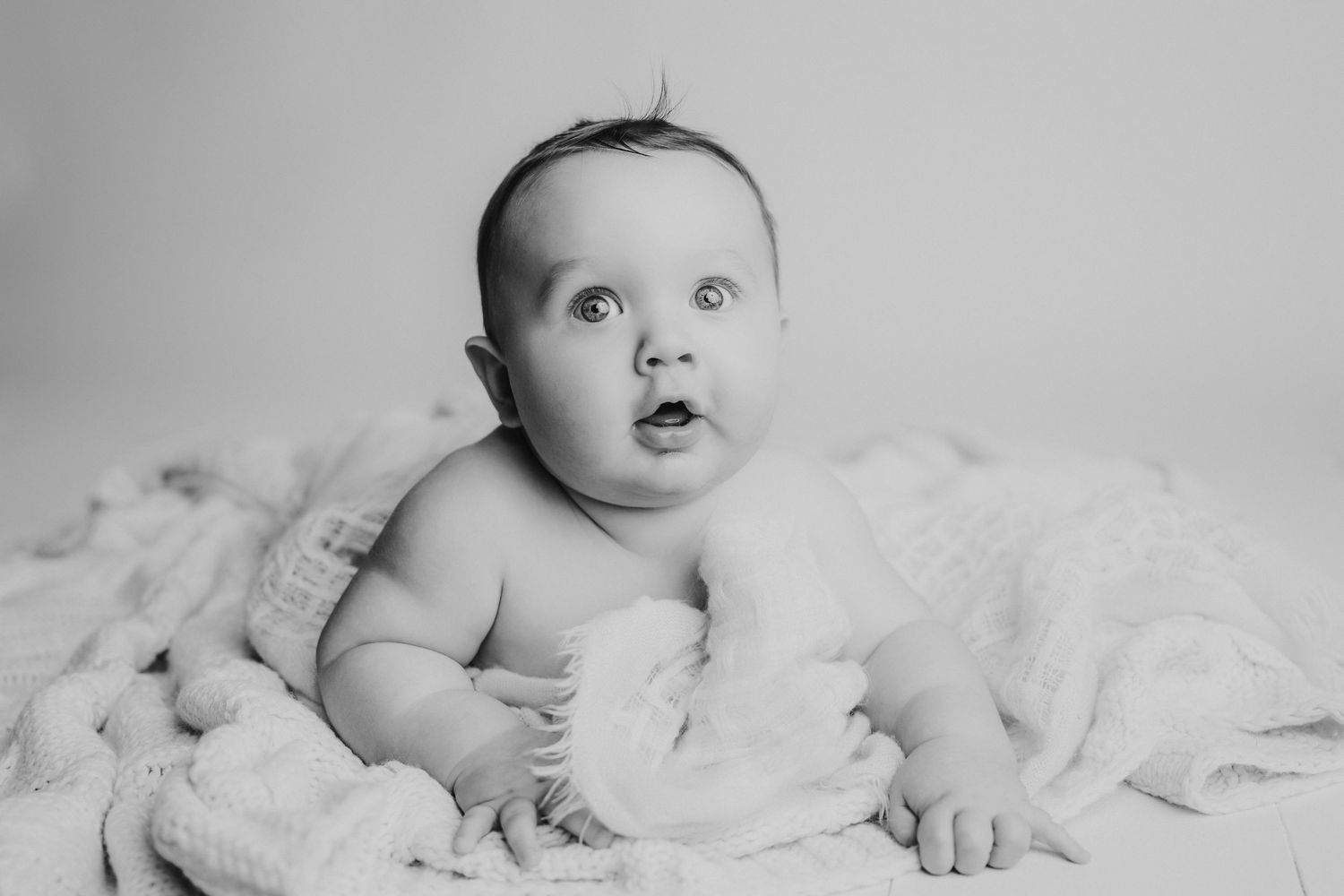 Black and white portrait of a baby lying on soft blankets with an expression of wonder.