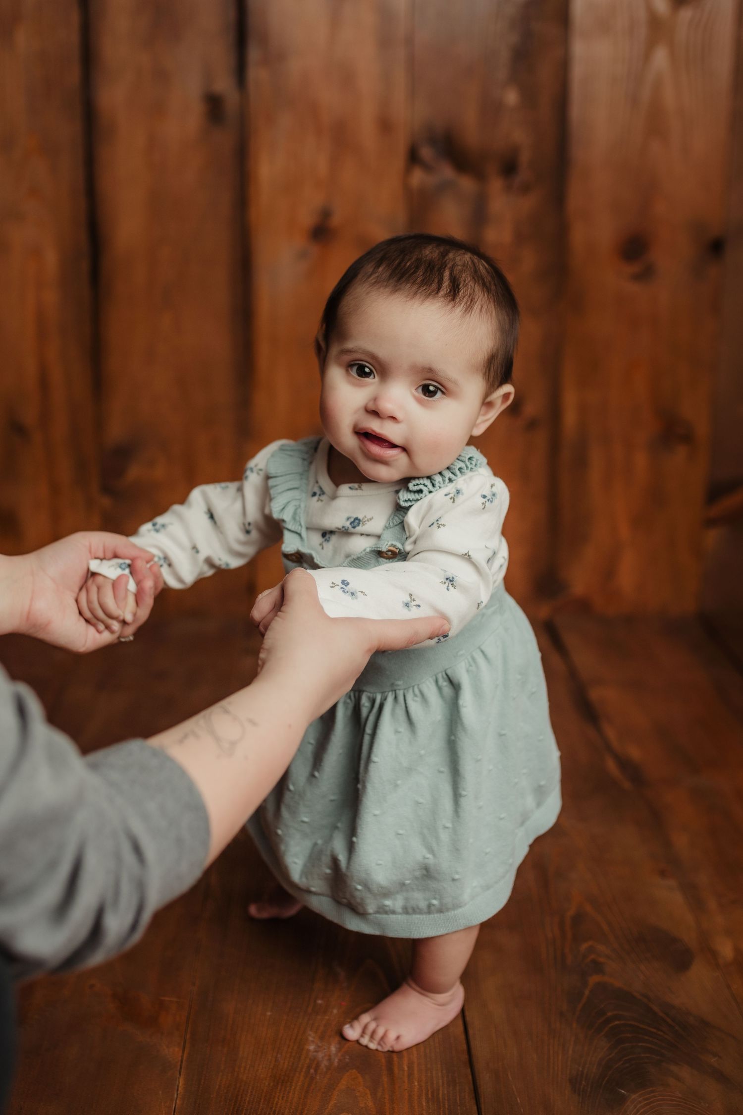 A baby in a sage green dress holds hands with someone while standing on hardwood floors.