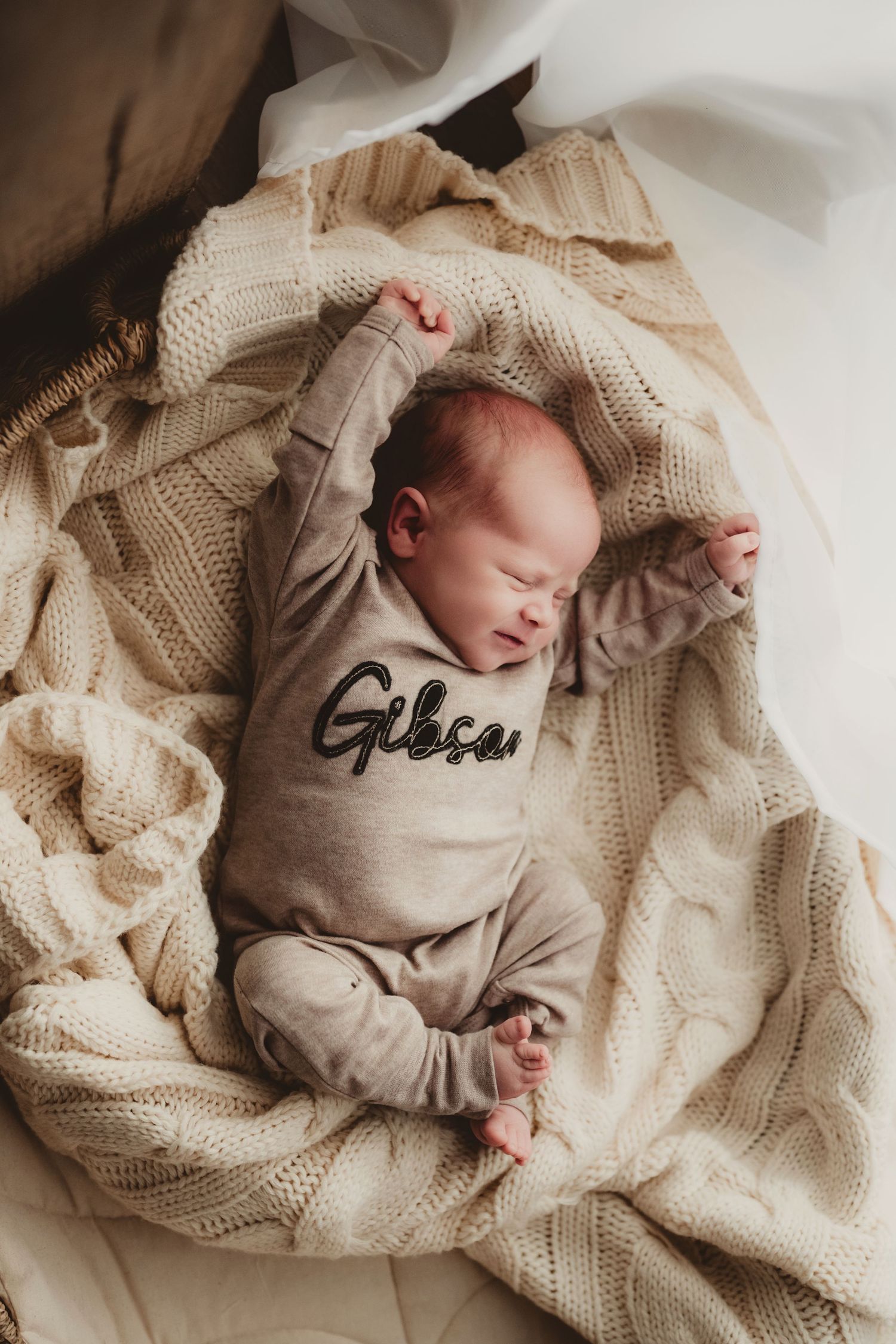 Sleeping newborn swaddled in cream knit blanket wearing light beige onesie on neutral backdrop.
