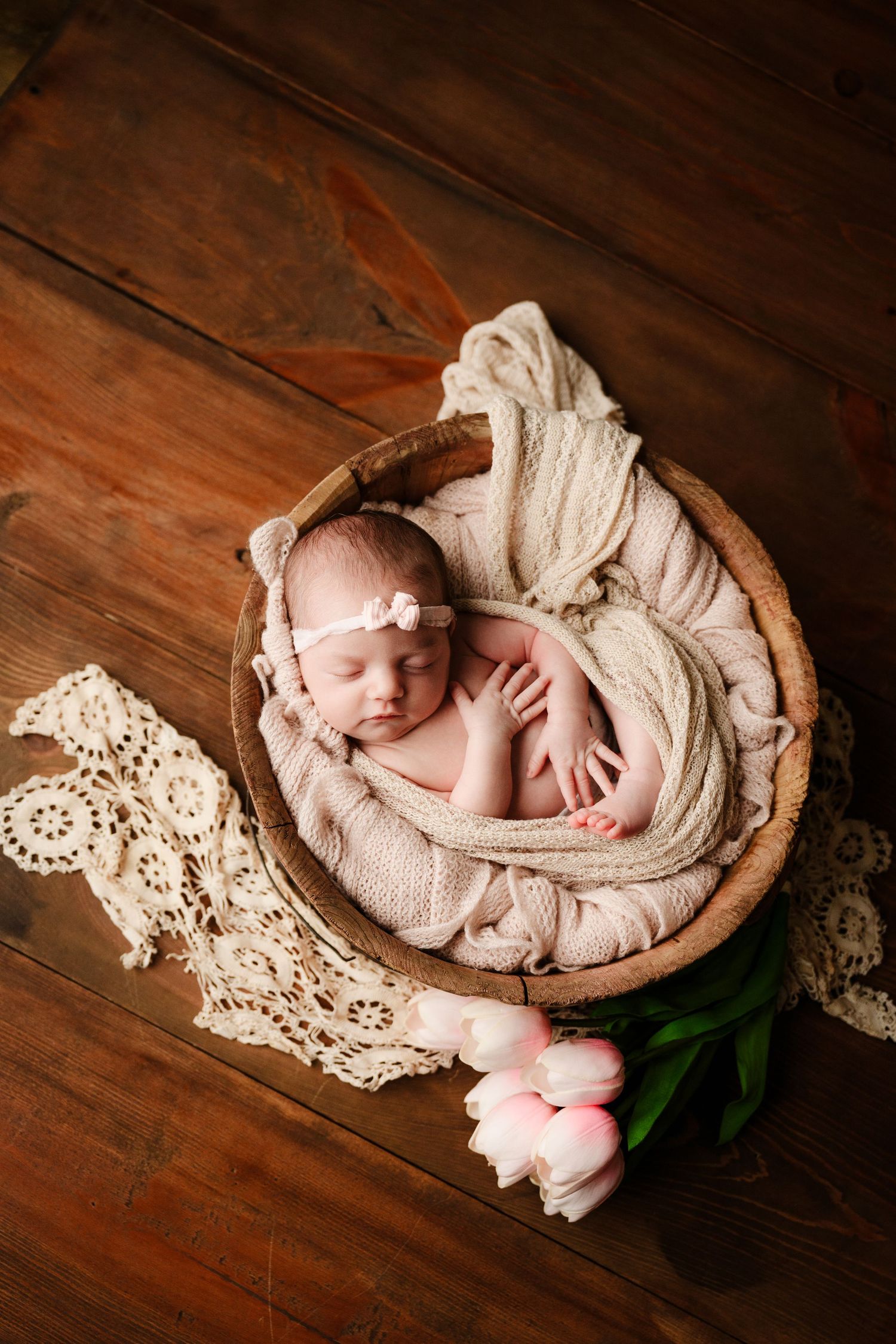 Newborn baby sleeping in wooden bowl with white wrap and pink tulips on wooden floor.