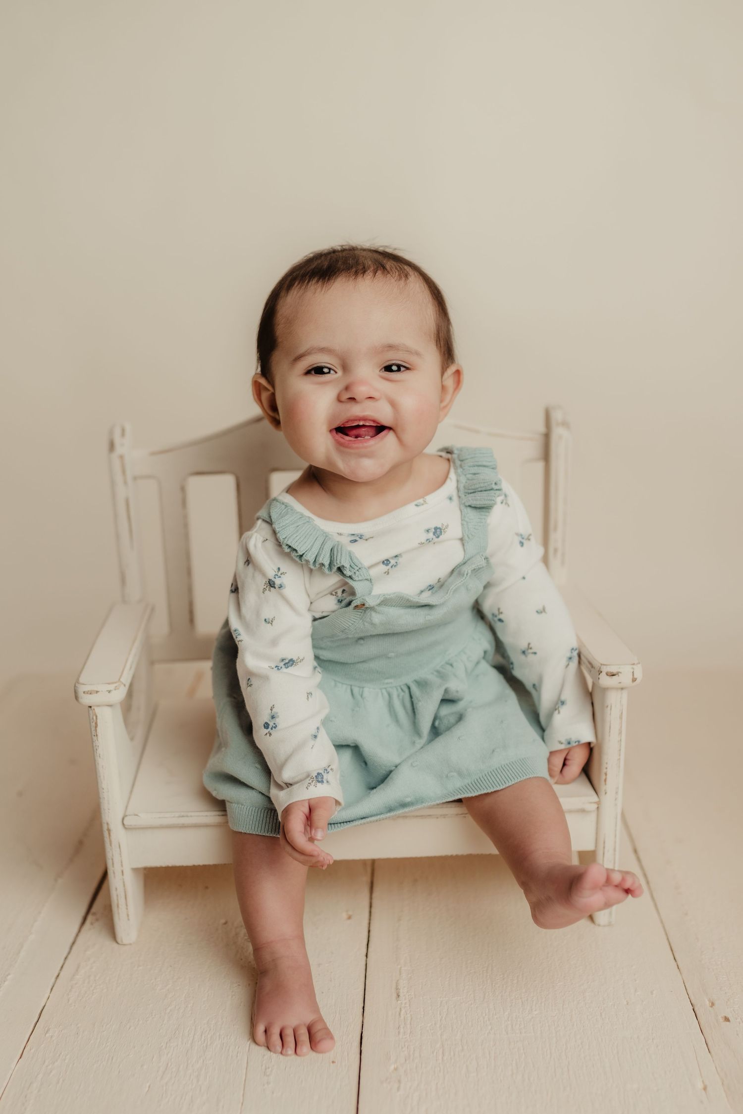 Happy baby in mint green dress sitting on white wooden chair.