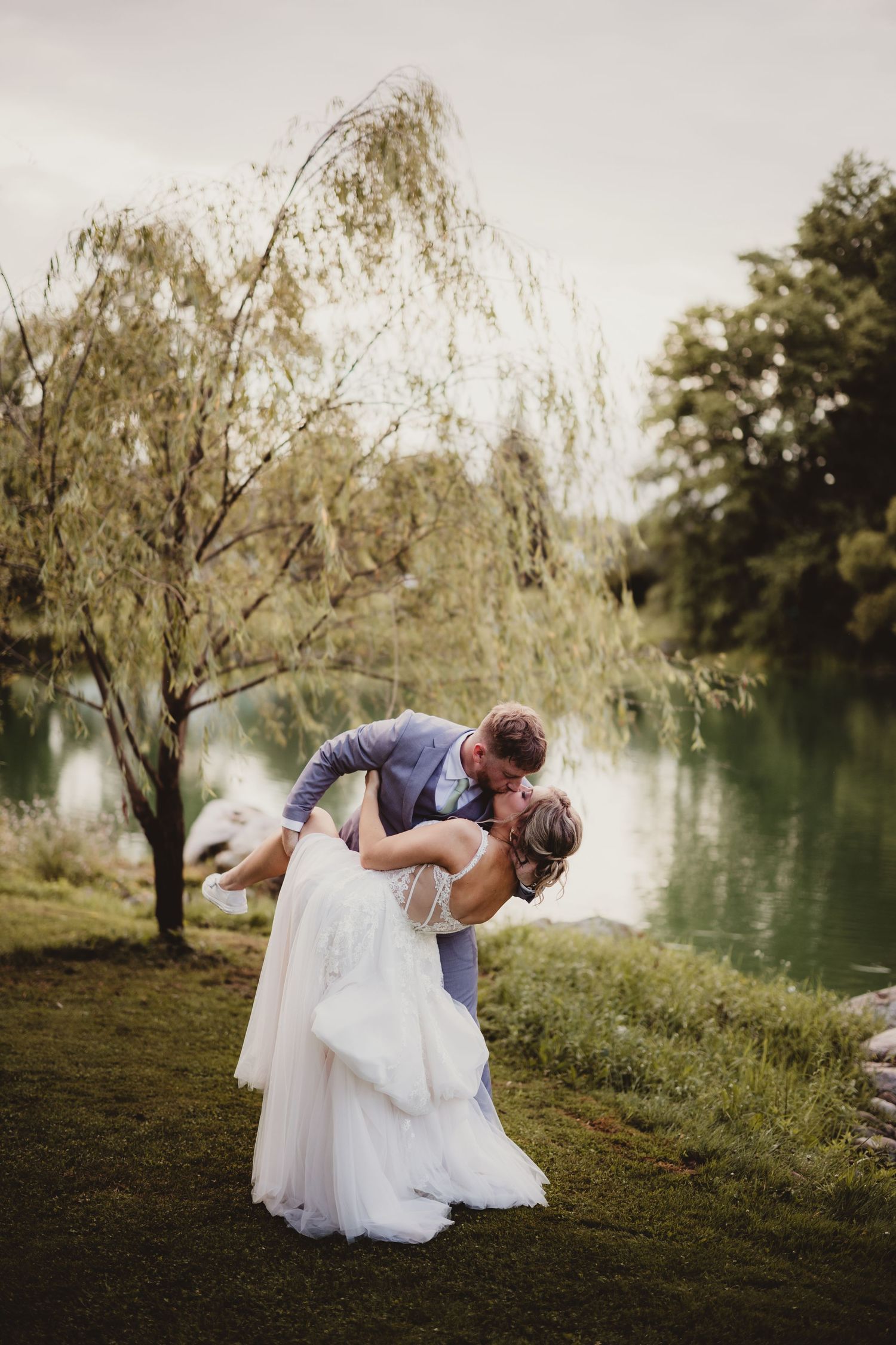 Romantic wedding couple share a passionate kiss at lakeside while groom dips bride at sunset near willow trees.