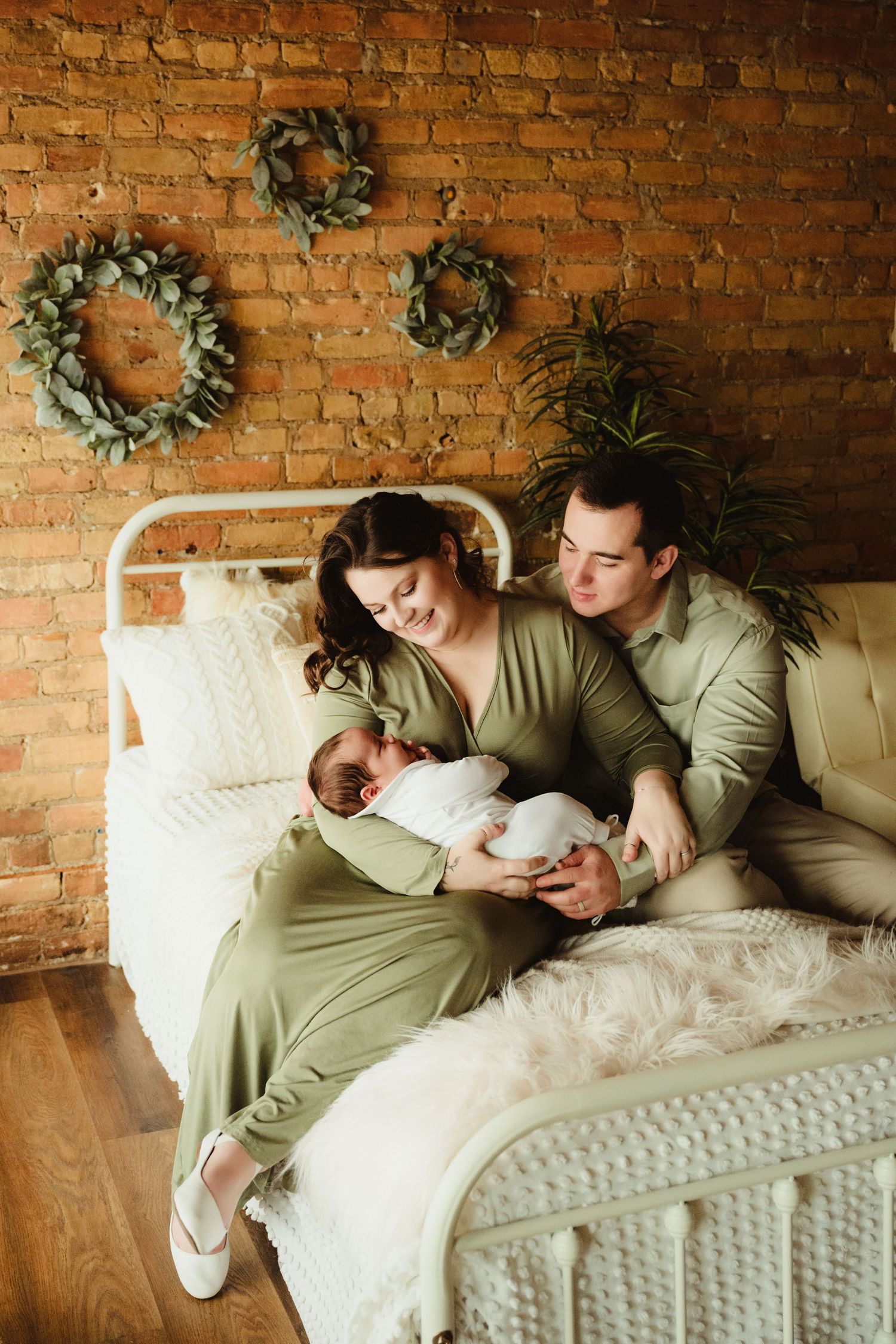 Couple in matching olive outfits resting on white bed with exposed brick wall and wreaths.