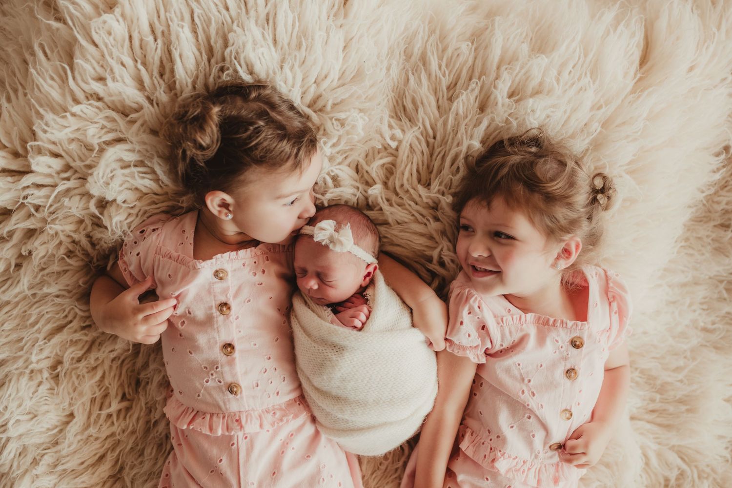 Three siblings in pink outfits lying together on fluffy rug.