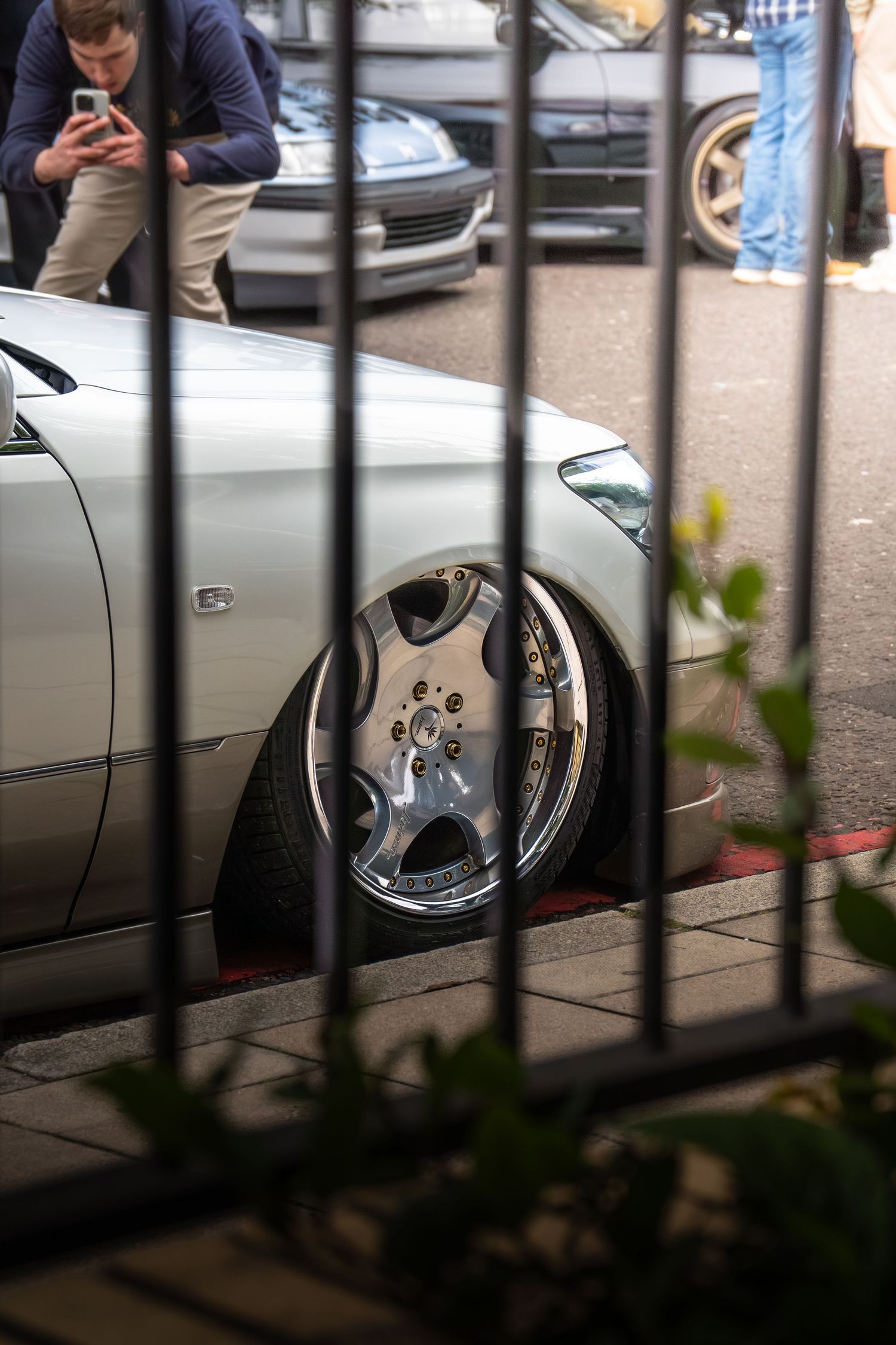 Lowered silver Mercedes with polished multi-spoke alloy wheels photographed through iron railings.