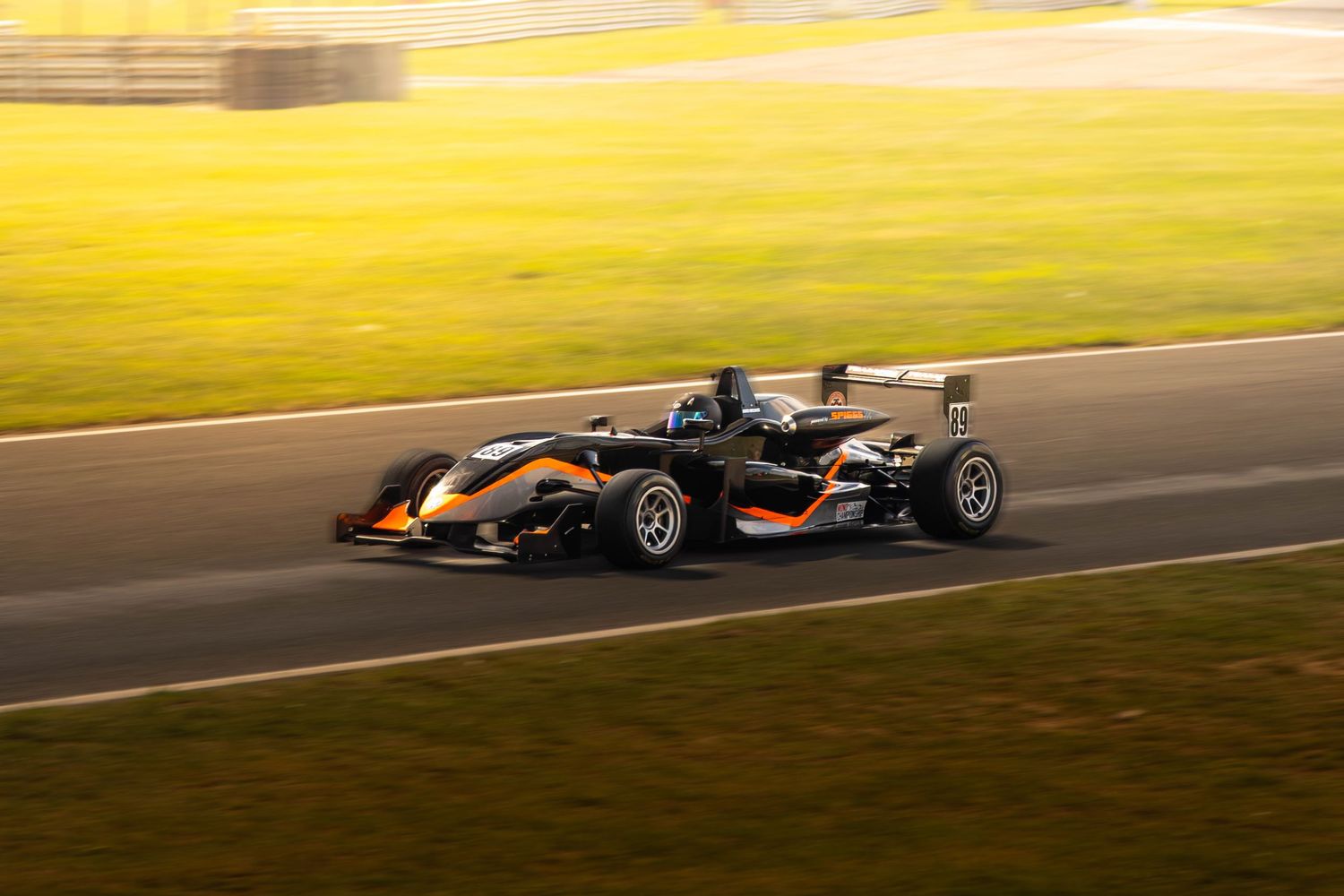 Black and orange Formula 3 racing car accelerates along a golden sunlit race circuit.