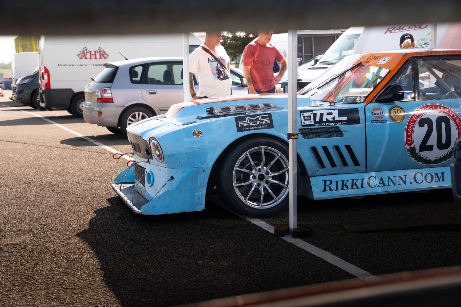Light blue Marcos racing car number 20 sponsored by RikkiCann.com in paddock area.