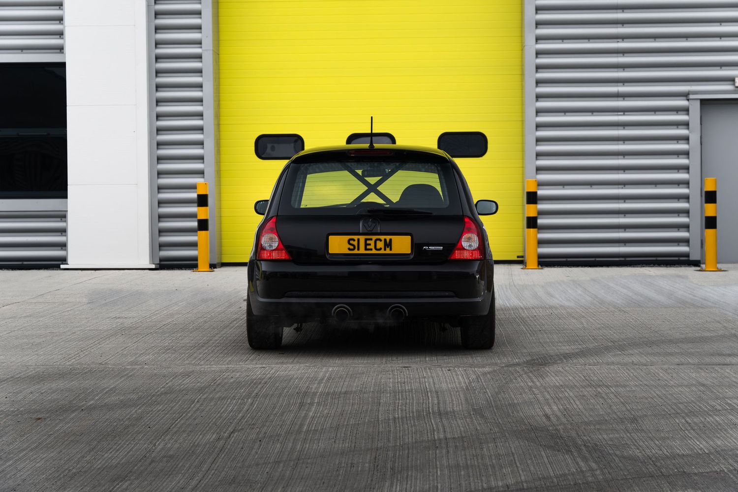 Close rear view of black Renault Clio S1ECM against a yellow and grey industrial roller shutter door.